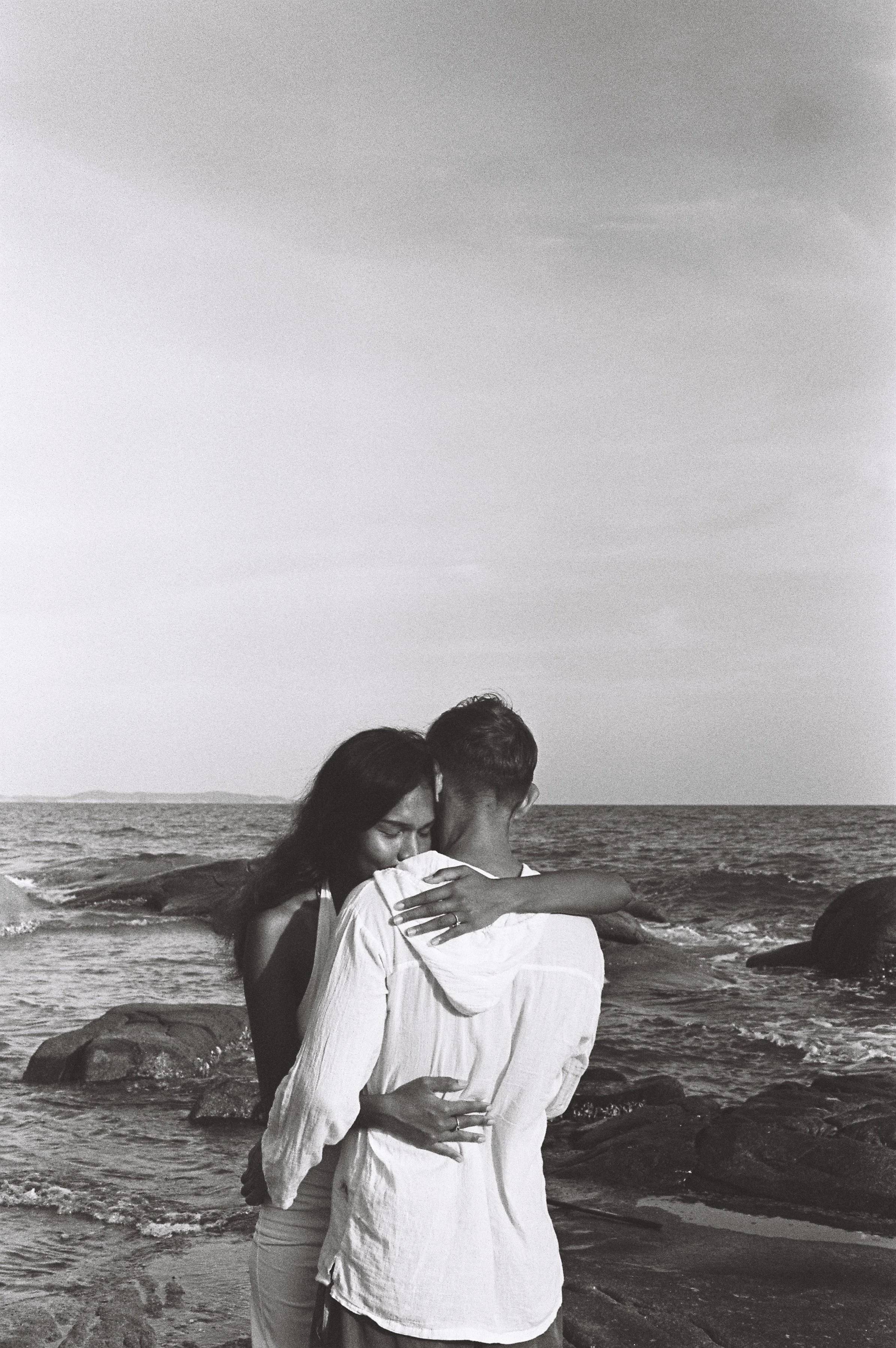 A black and white photo of a couple hugging on a beach. The woman has long dark hair and is leaning her head on the man's shoulder, who has short hair. They are embracing near the water with rocks and the ocean in the background.