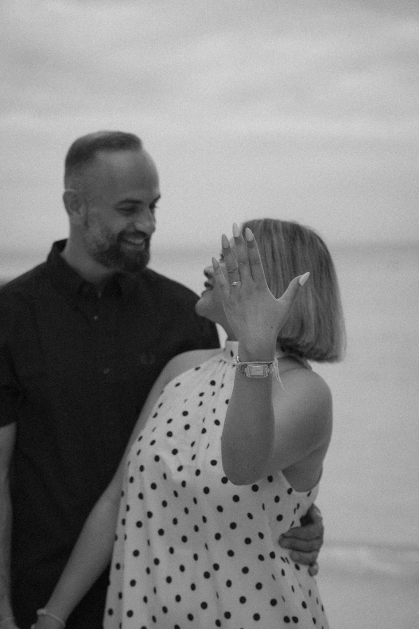 A man and woman are dancing closely together on a beach in a black-and-white photo. The man has a beard and short hair, smiling while looking at the woman. The woman, facing away, has long hair and is wearing a polka dot dress, with her hand raised a