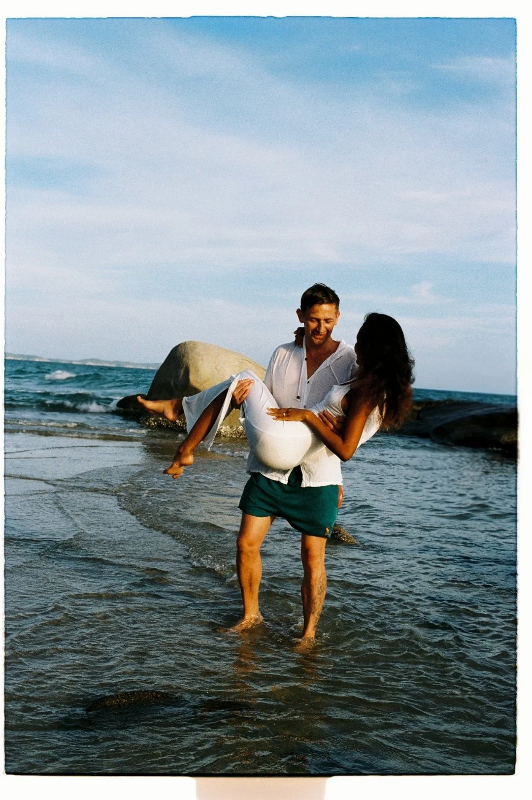 A man is holding a woman in his arms at the beach, both smiling, with rocks and the ocean in the background.