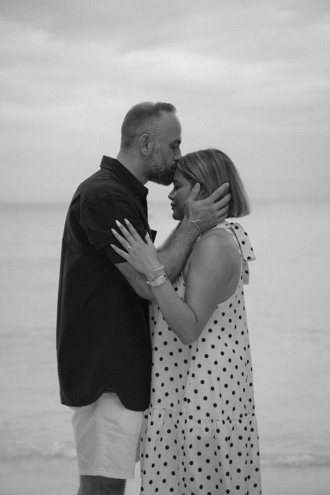 A man with a beard and a woman with short hair are holding each other closely on the beach. The man is wearing a black shirt, and the woman is wearing a polka dot dress. He is gently holding her face, and she is holding his arms. They are sharing an 