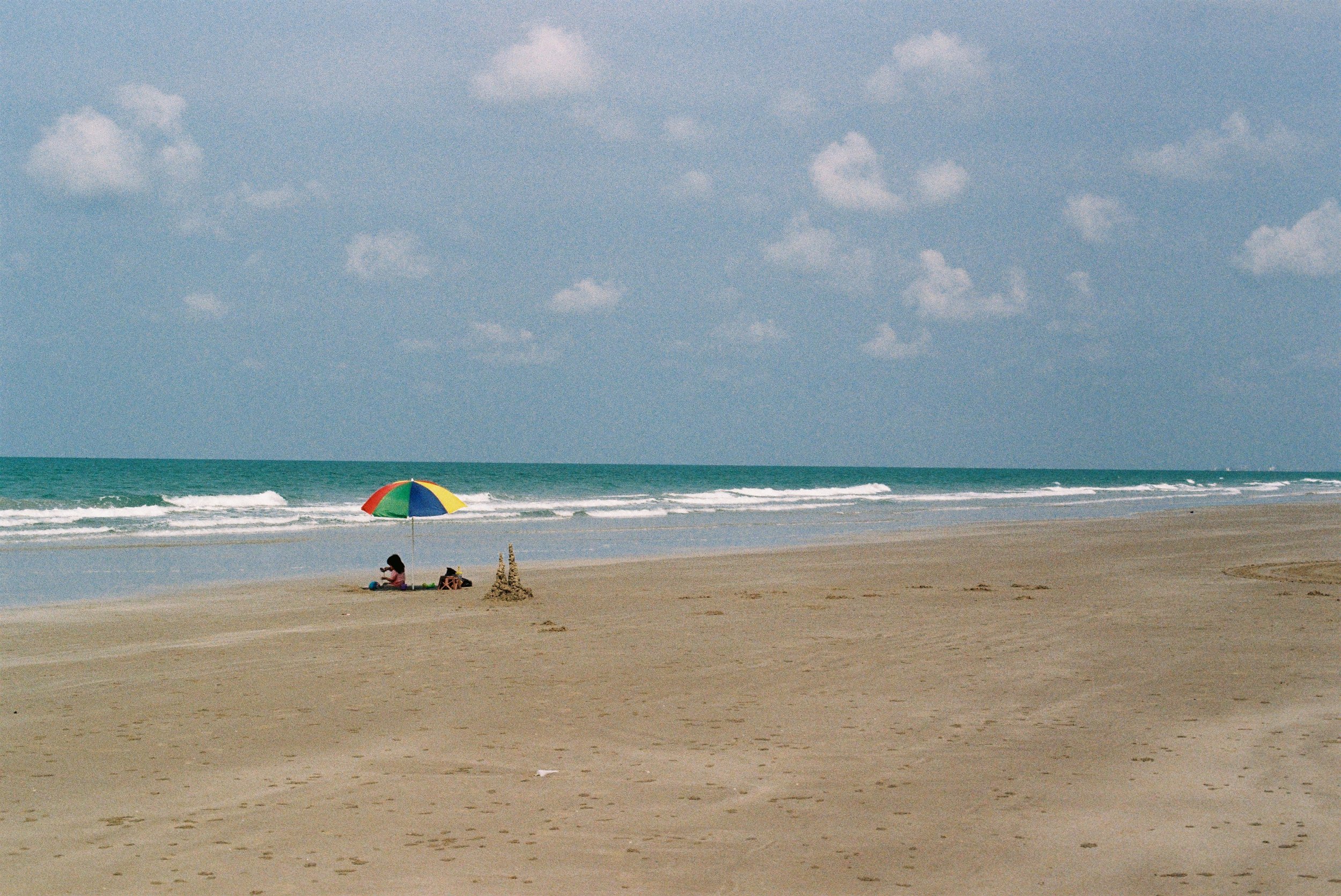 Person sitting under a multicolored beach umbrella on a deserted sandy beach with ocean waves and a partly cloudy sky.