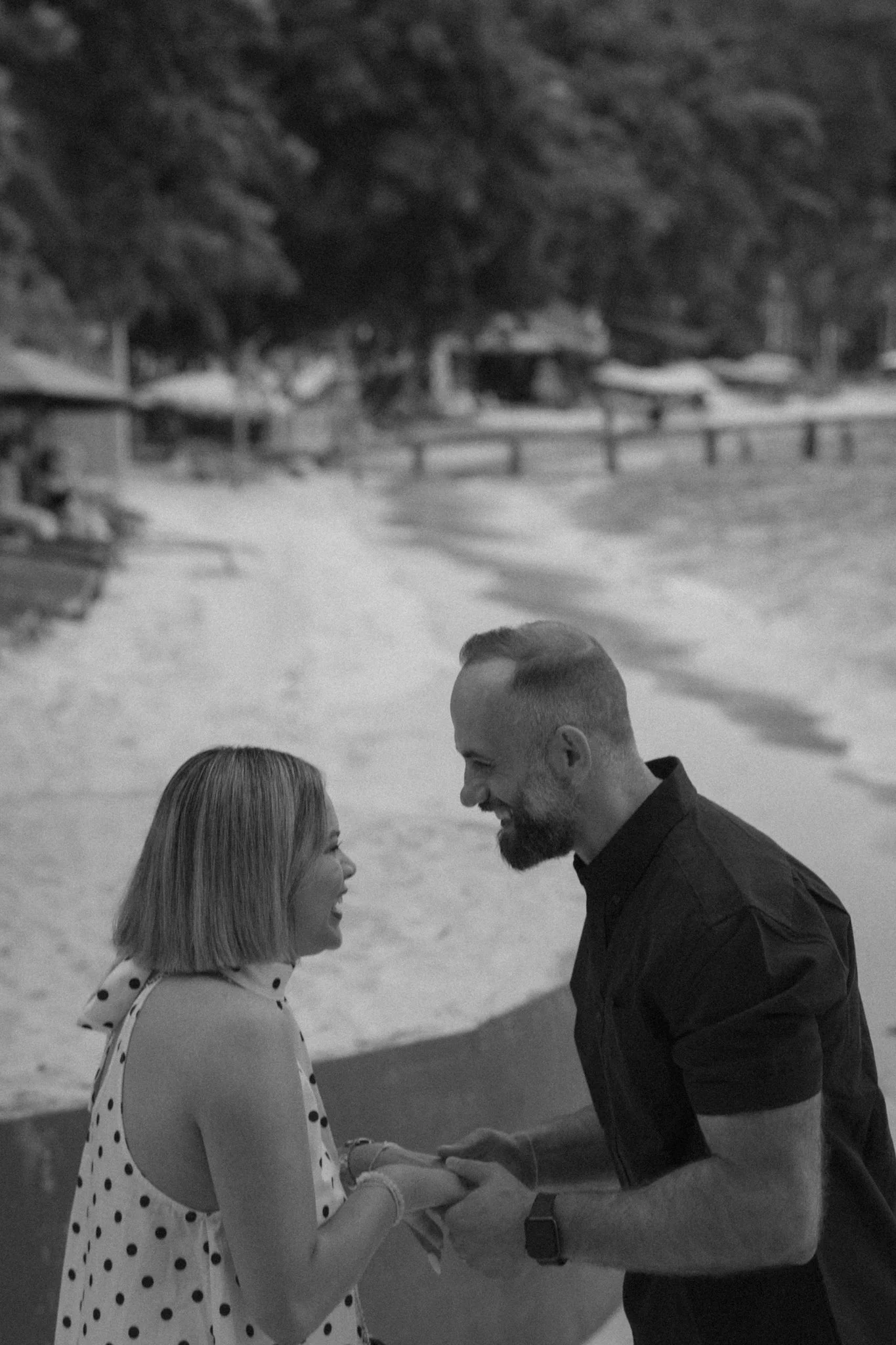 A man and woman are facing each other outdoors near a body of water, smiling and holding hands. The woman is wearing a polka dot dress, and the man is dressed in a dark shirt. Trees and a fence are visible in the background.