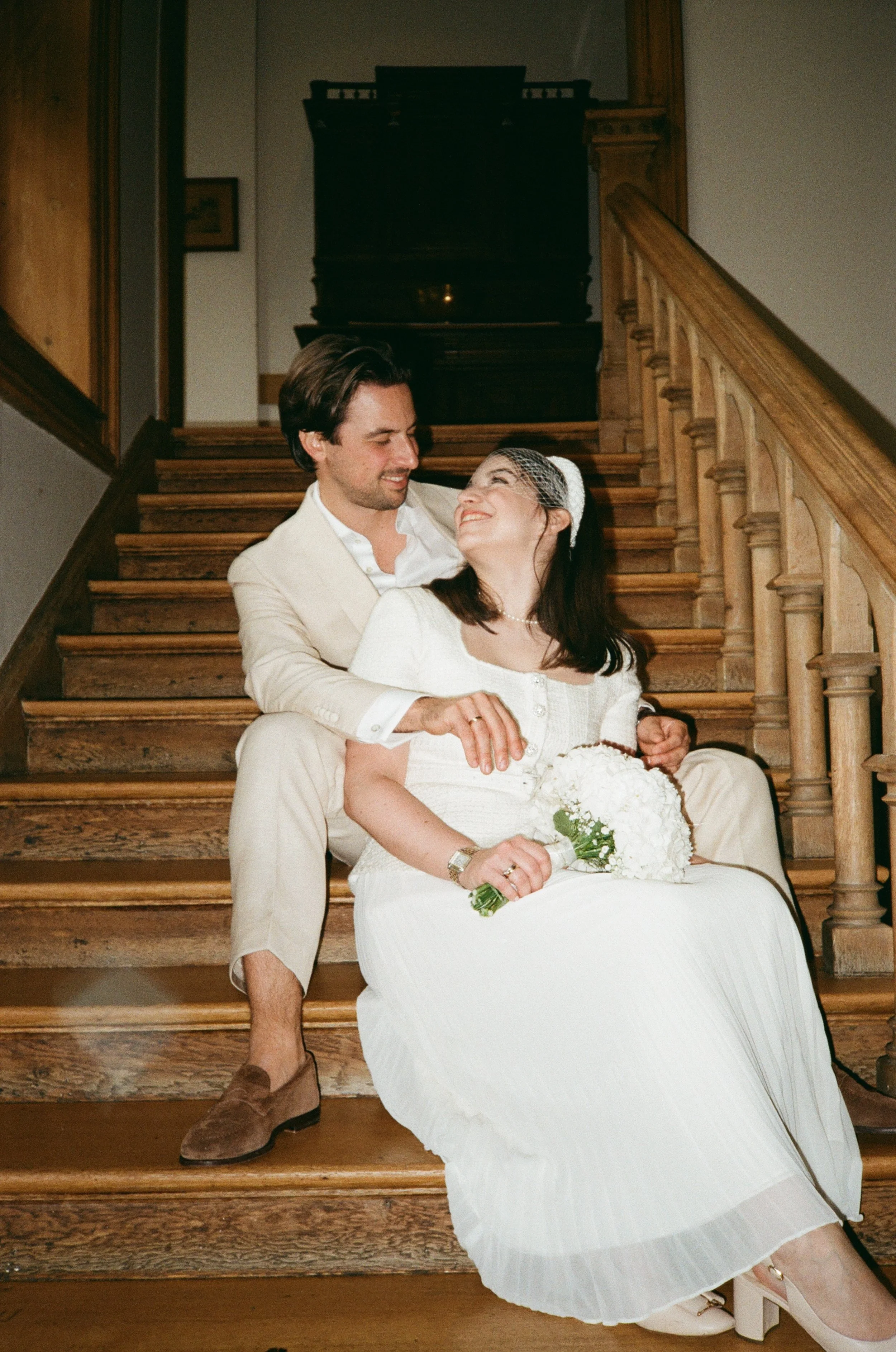 A man and woman sitting on wooden stairs, smiling at each other. The woman is holding a bouquet of white flowers, wearing a white dress with a pearl necklace and a birdcage veil. The man is dressed in a light-colored suit with brown shoes. The backgr