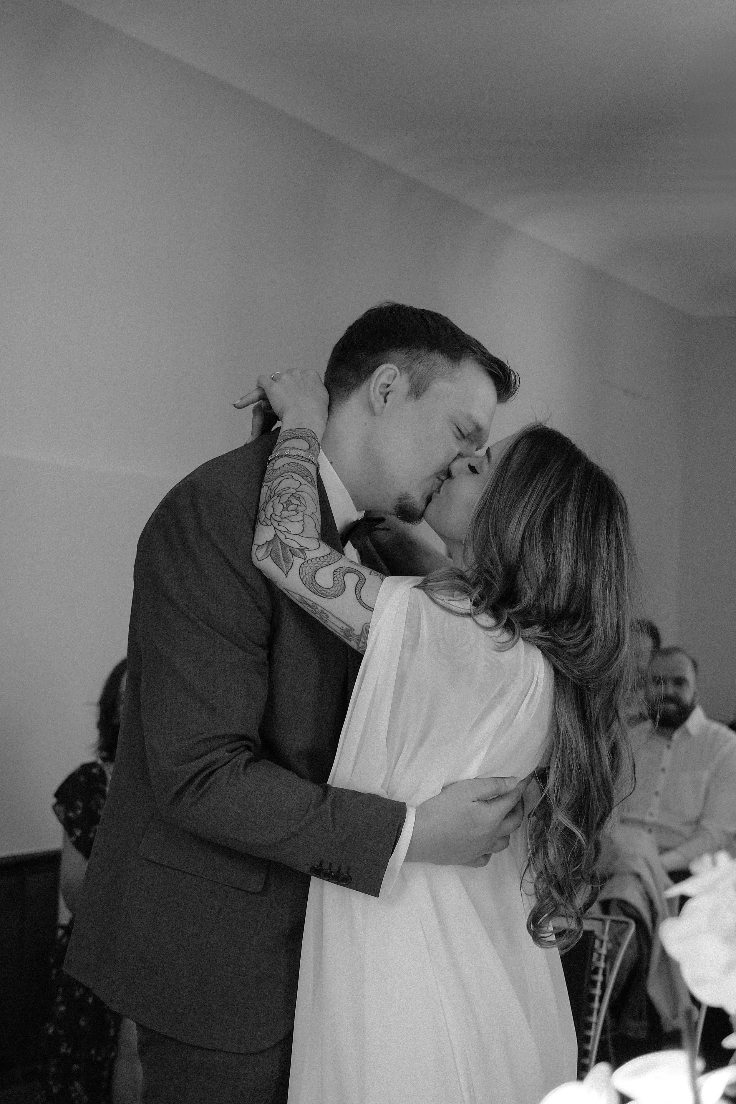 A black-and-white photograph of a couple sharing a kiss, with the man in a suit and the woman in a light-colored dress, at a gathering or celebration.