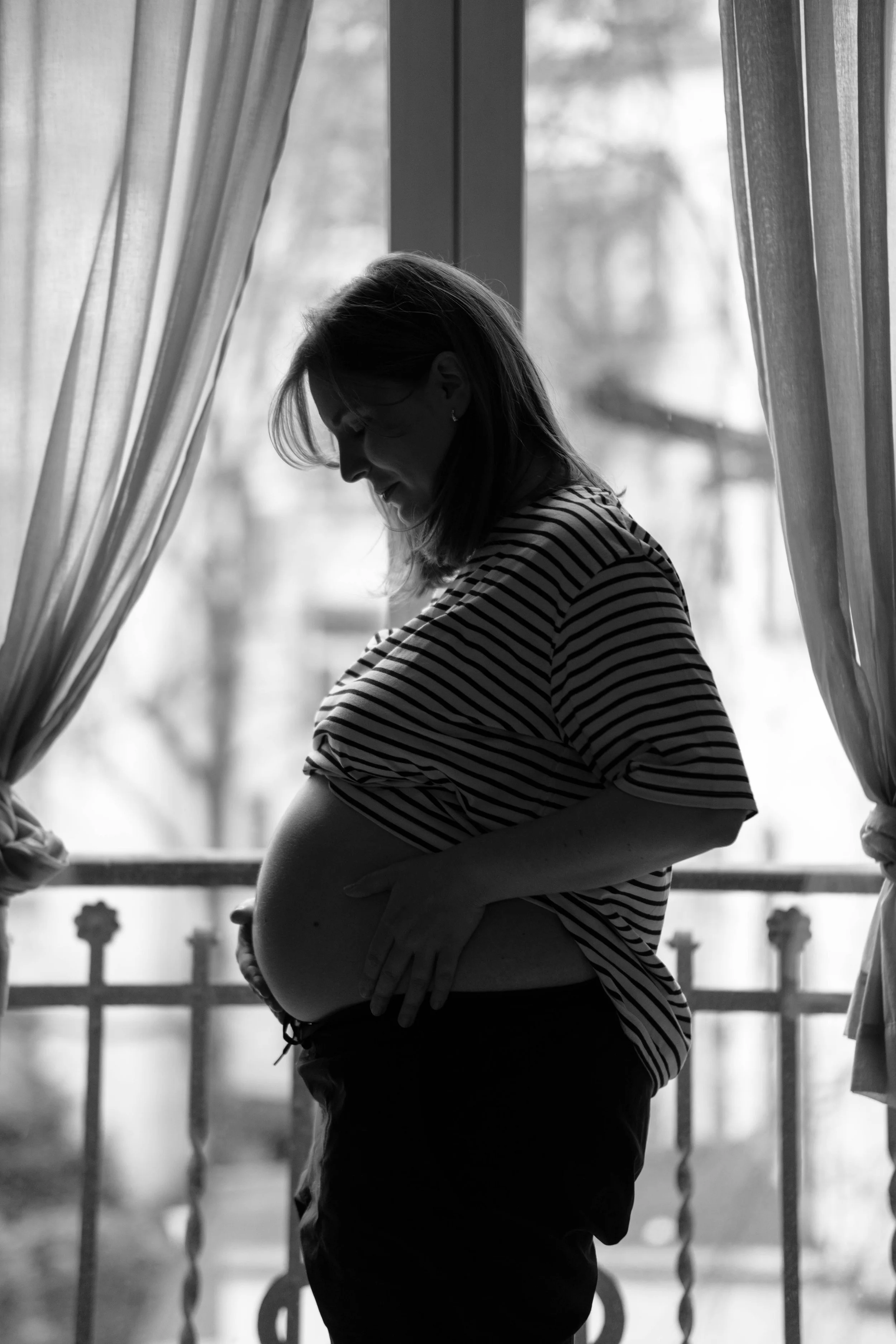 Silhouette of a pregnant woman standing by a window, with her hands on her belly and a gentle expression, surrounded by curtains.