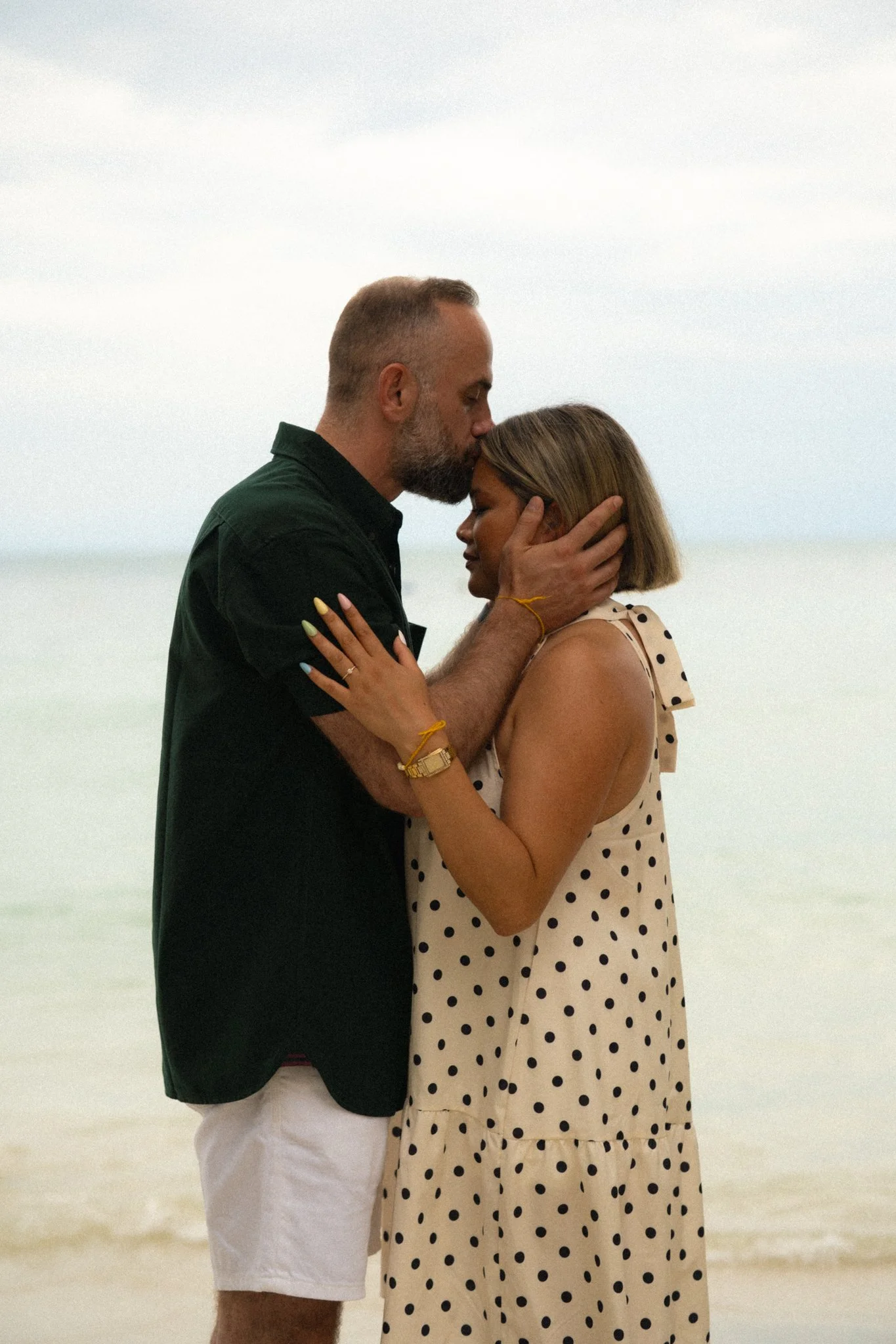 A man and woman embrace on the beach. The man is kissing the woman on her forehead while holding her face, and she is holding his hand. The man is wearing a black shirt and white shorts, and the woman is wearing a polka dot dress. The background is a