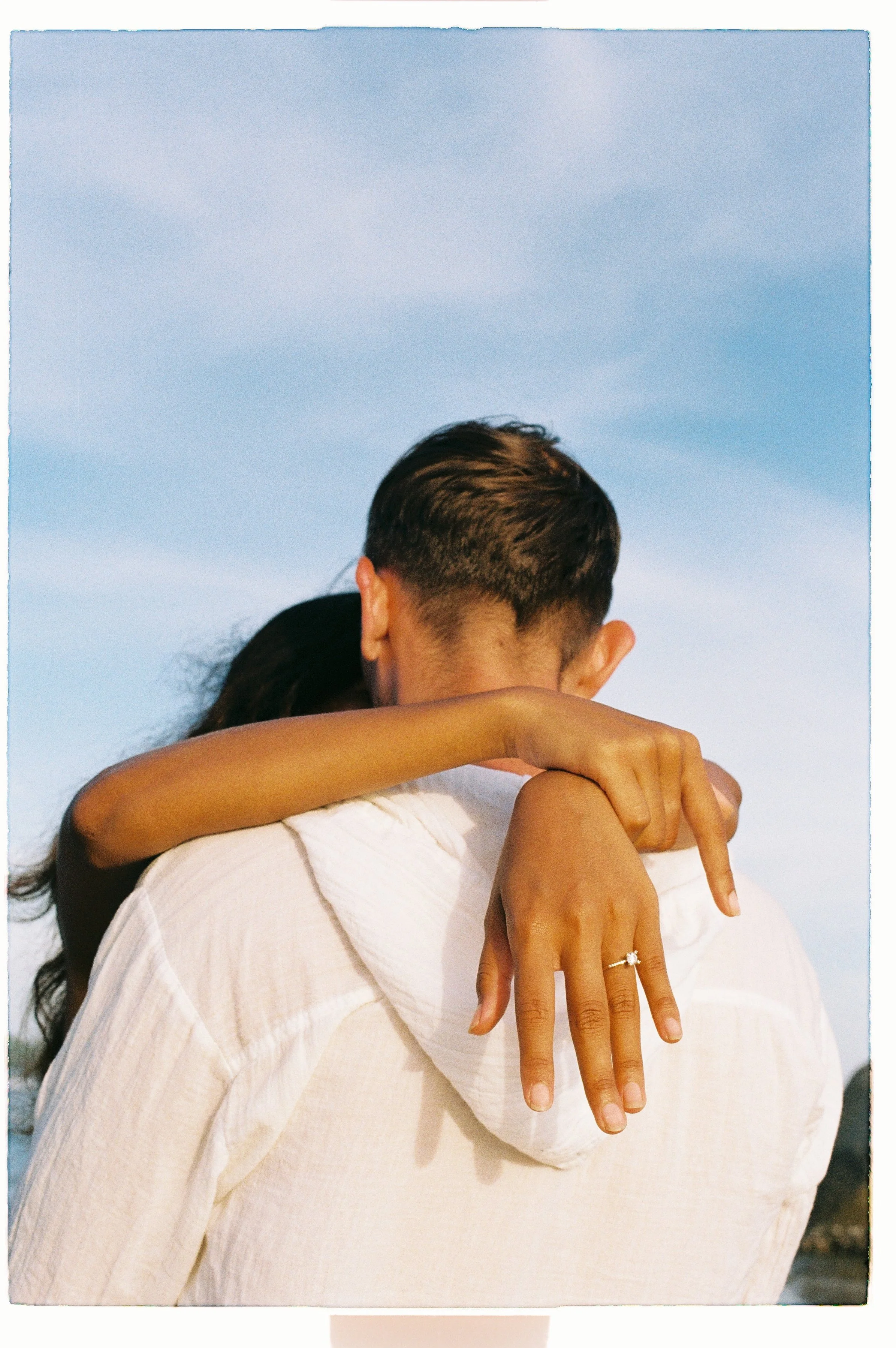 A close-up of a couple embracing, with the woman's hand displaying a diamond ring and her arm draped over the man's shoulder against a blue sky background.