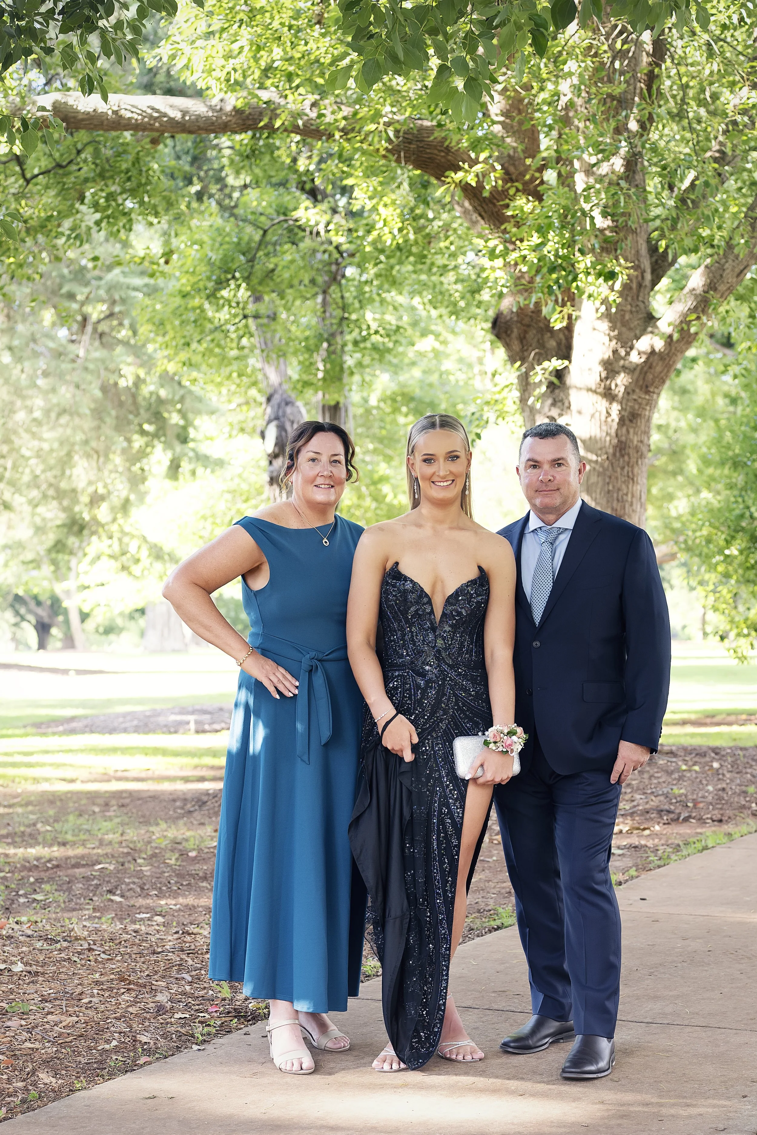 Three people dressed formally standing on a park path with green trees in the background, smiling at the camera.