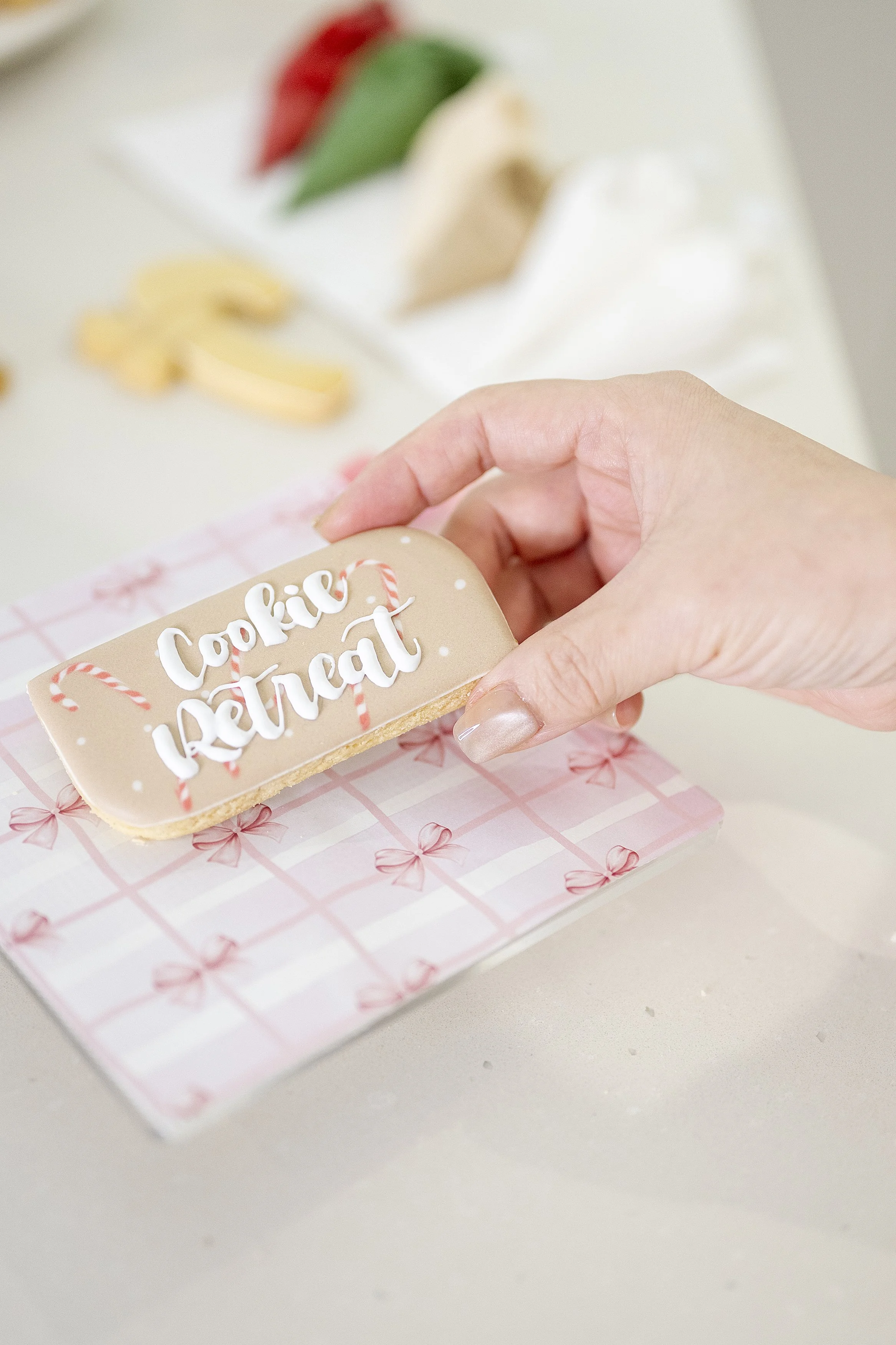 Close-up of a hand holding a decorated sugar cookie with the words "Cookies ReaT" written on it, on a pink and white checkered gift tag with pink bows. In the background are more cookies on a white surface, including a red, green, and beige cookie.