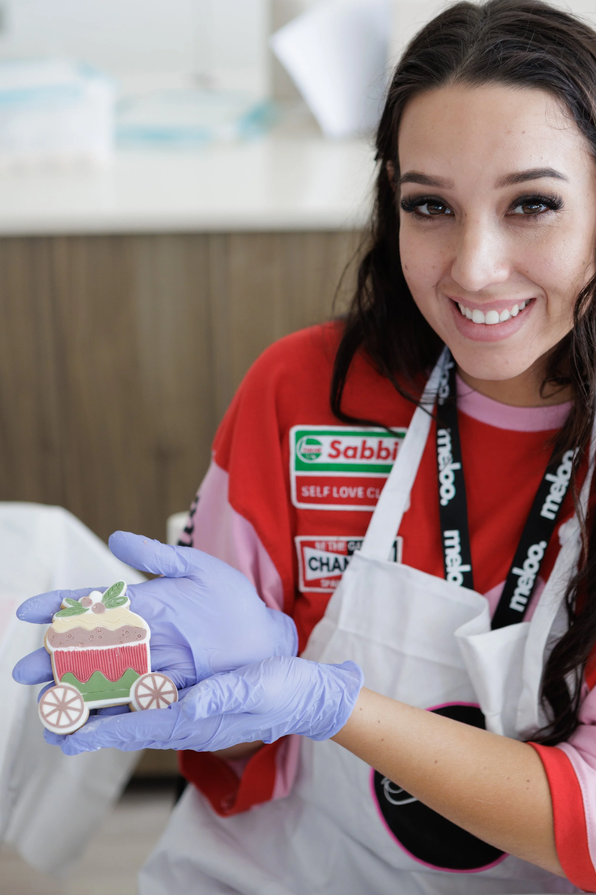 A woman smiling and wearing gloves, holding a decorated cookie shaped like a Christmas present on wheels, in what appears to be a kitchen or bakery setting.