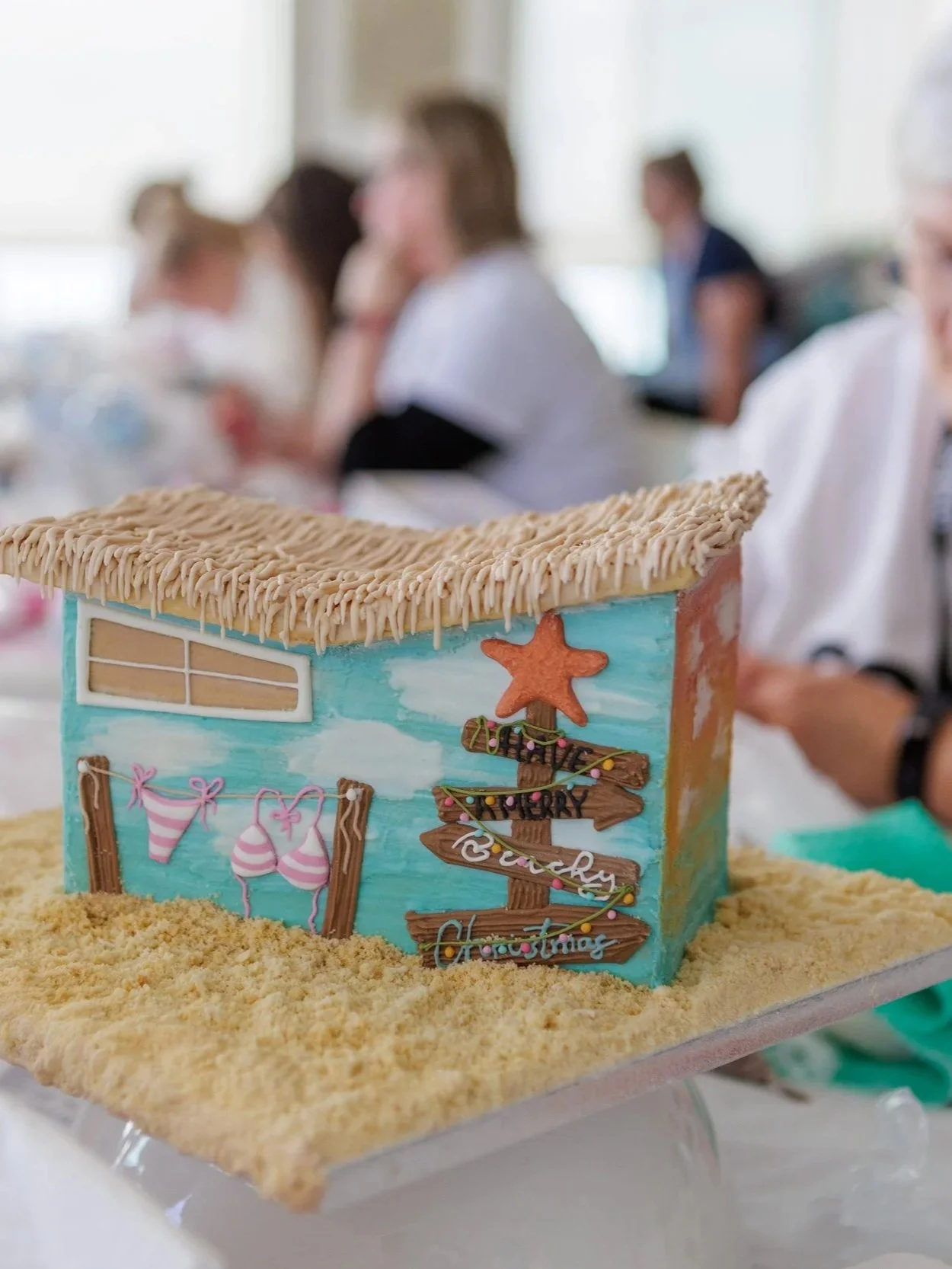 A rectangular cake decorated with a beach scene, including a starfish, wooden signpost, and bikinis on a laundry line, set on a sandy surface, with people blurred in the background.