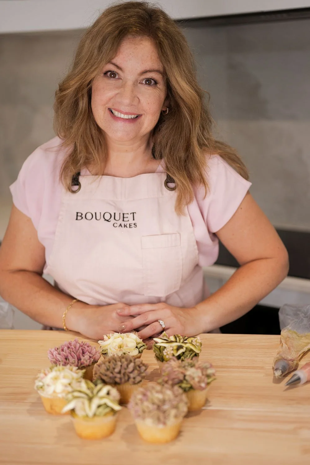 Woman with brown hair wearing pink apron stands behind her cupcake creations in a creative session photoshoot