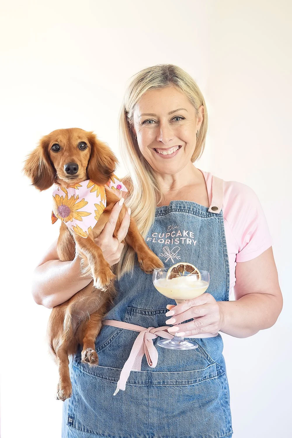 Blonde woman in a blue apron holds a tan dachshund and a cocktail in a branding shoot