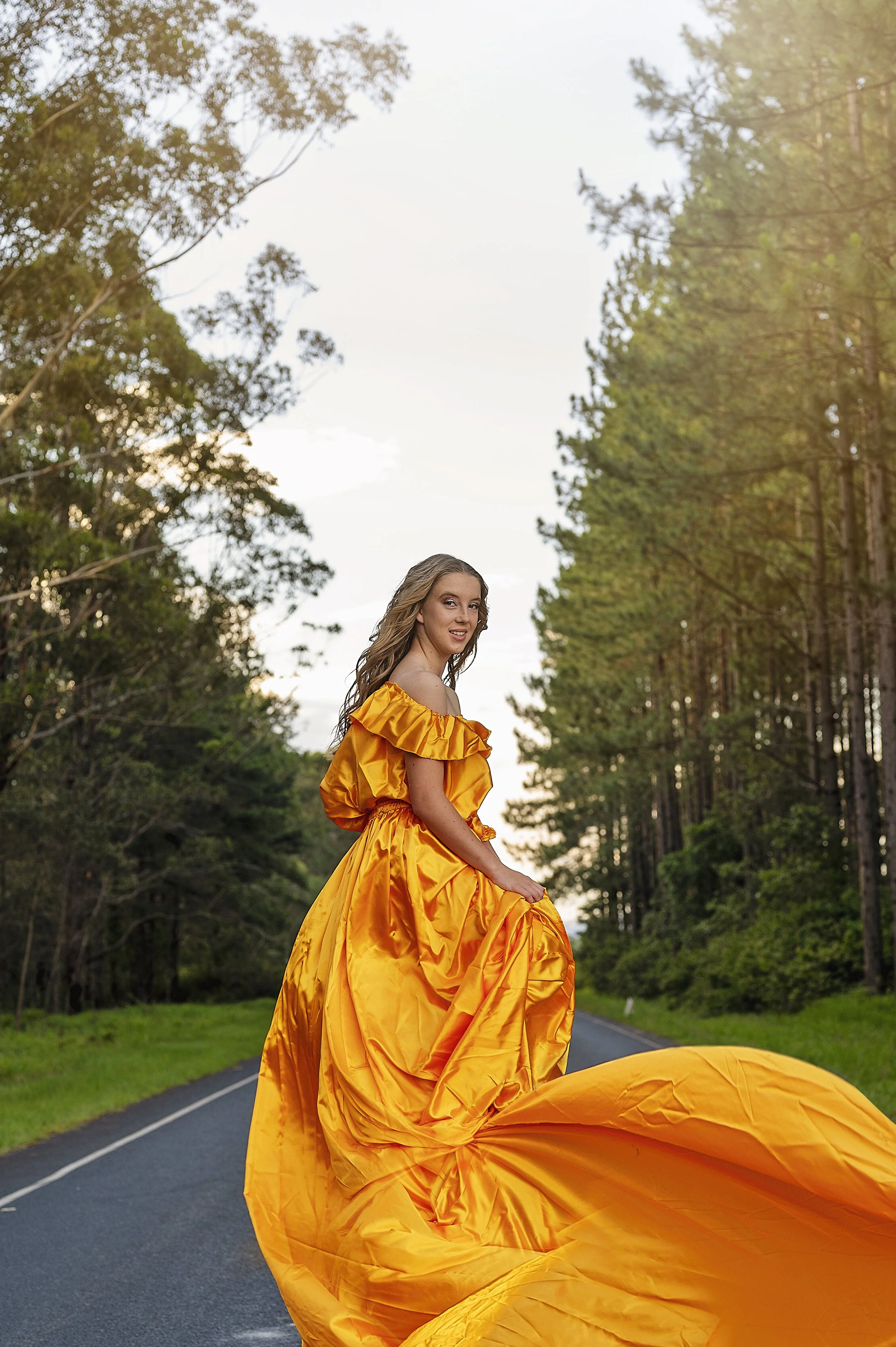 A woman in a bright yellow, elaborate gown standing on a road surrounded by trees, during sunset.