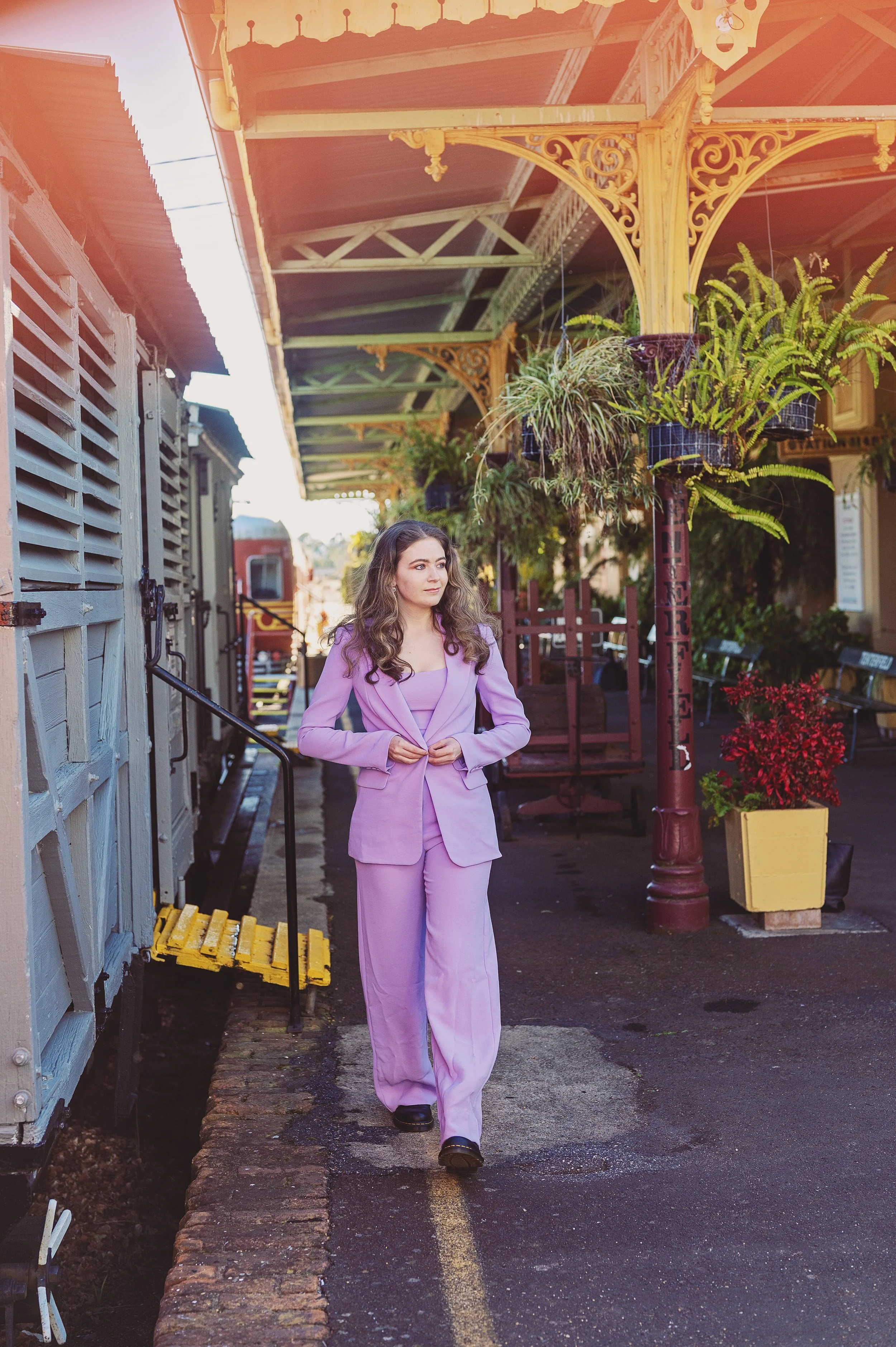 A woman in a pink suit walking along a historic train station platform with potted plants and ornate ironwork.