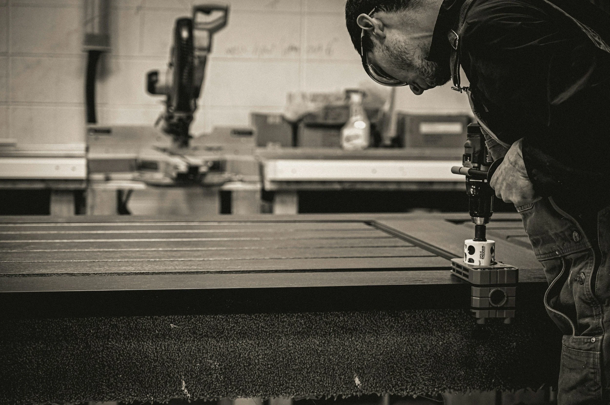 A person wearing glasses and a jacket is working with a calibrated device on a textured surface, likely in a workshop or manufacturing environment. The background contains various tools and materials.