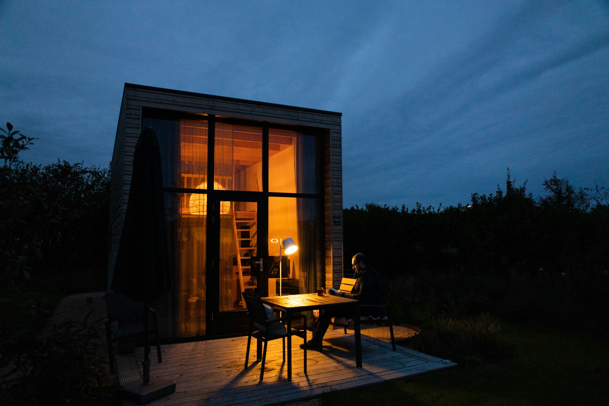A modern tiny house with large glass windows and wooden exterior, illuminated from within during evening, surrounded by bushes and trees, with a person sitting at a desk outside.