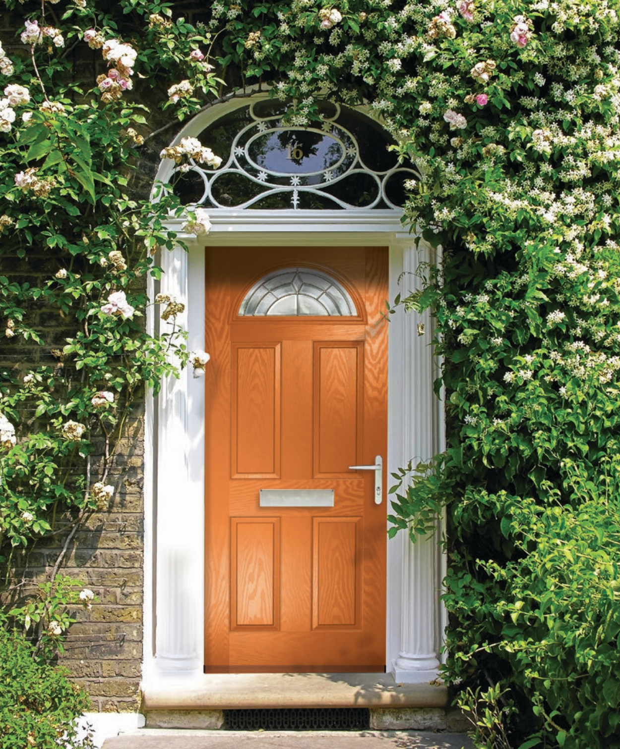 A wooden front door with a small window at the top, surrounded by white trim, set in a brick wall with lush green vines and white flowers growing around it.