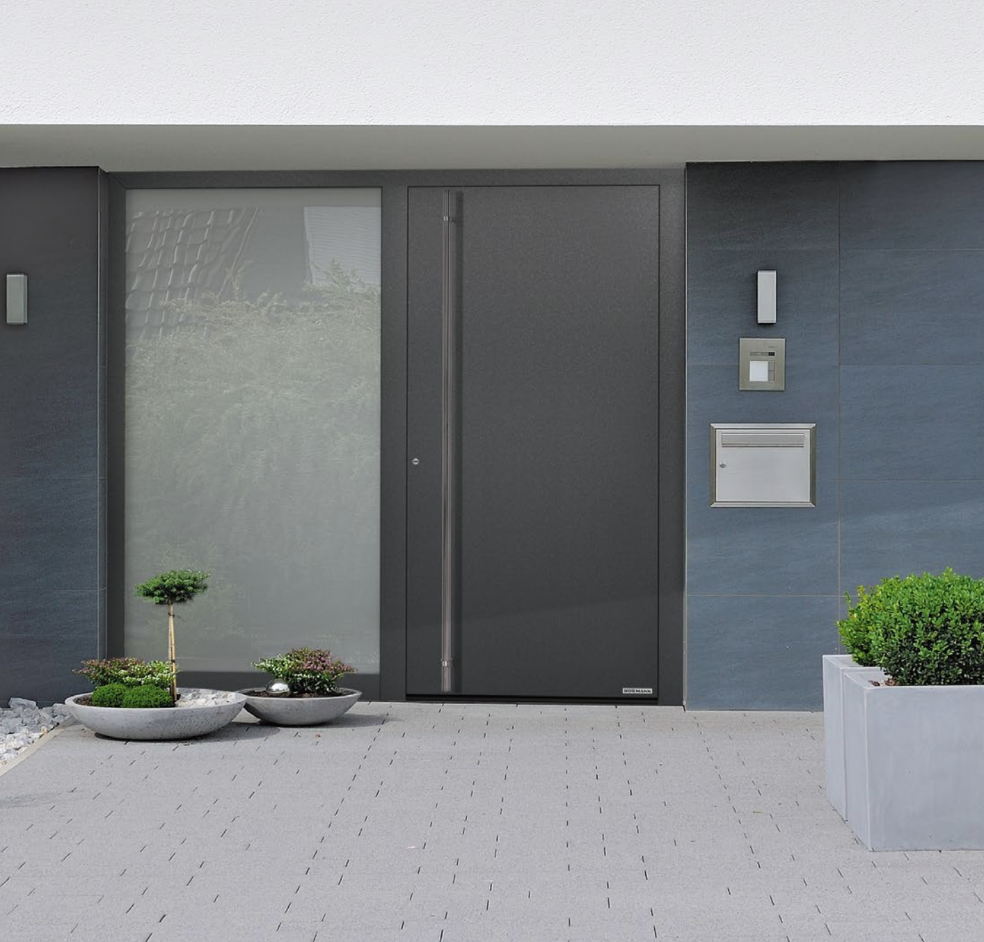 Modern building entrance with gray door, small trees and bushes in planters, and a gray tiled wall.