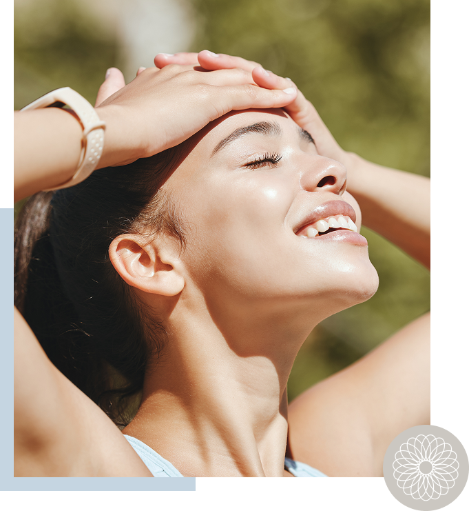 Close-up of a smiling woman outdoors with eyes closed, hands on her forehead, sunny weather.
