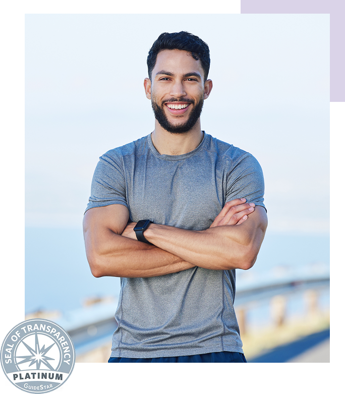 A smiling man with a beard and short dark hair, wearing a gray athletic shirt and a watch, standing outdoors near a coastal area with a bridge in the background.