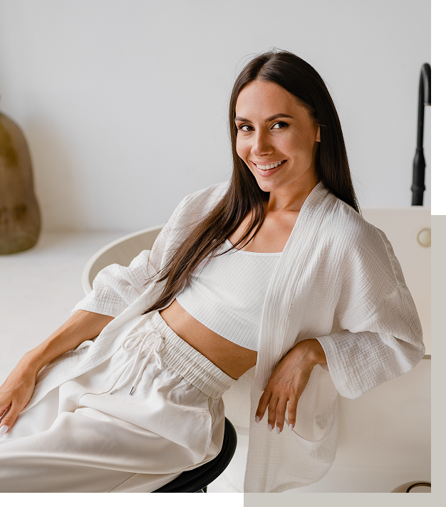 A woman sitting on a modern chair in a white room, smiling at the camera, wearing a white crop top, a white robe, and matching white pants.