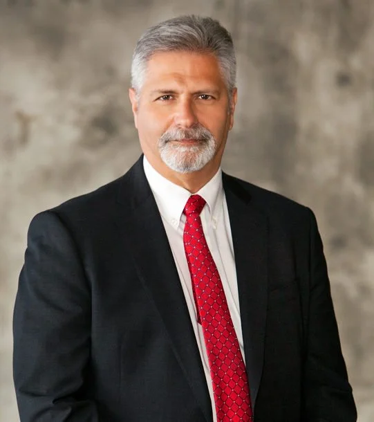 A mature man with gray hair and a beard, wearing a dark suit, white shirt, and red patterned tie, standing against a neutral background.