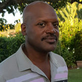 A man with a shaved head wearing a light-colored, collared shirt outdoors with greenery and trees in the background.