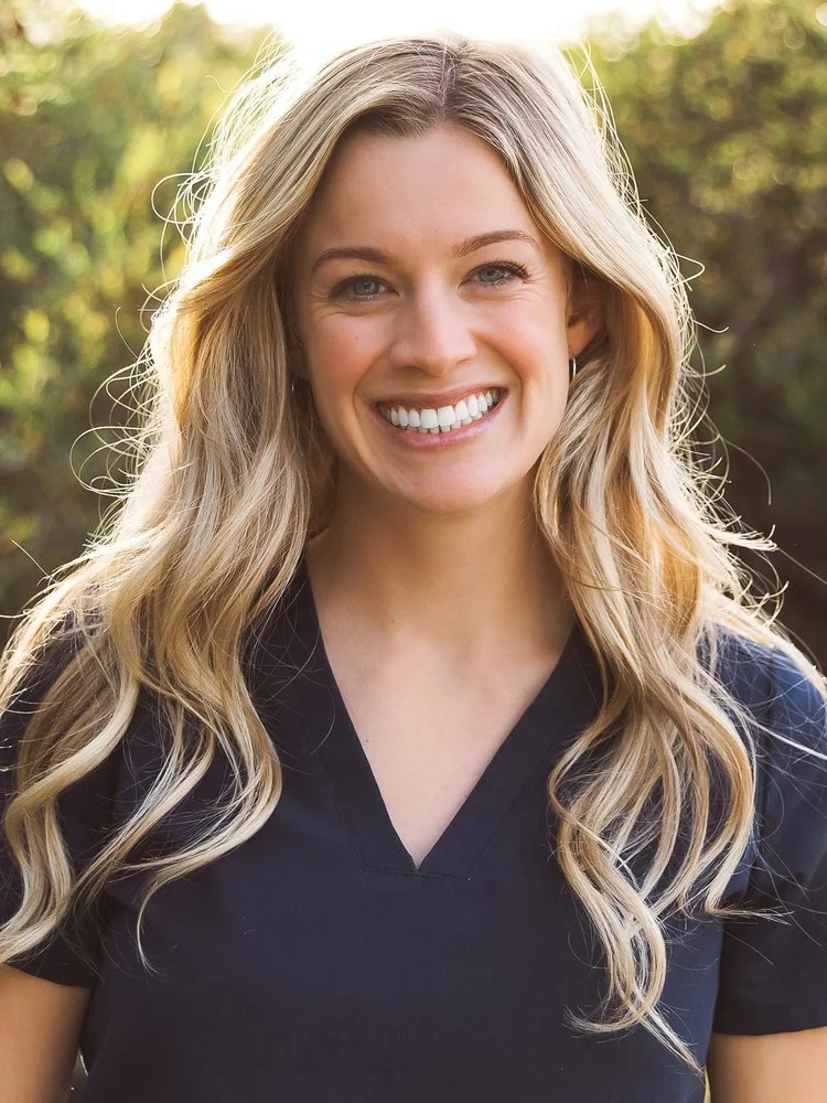 Portrait of a young woman with long blonde wavy hair, smiling, wearing a black top outdoors with blurred greenery background.