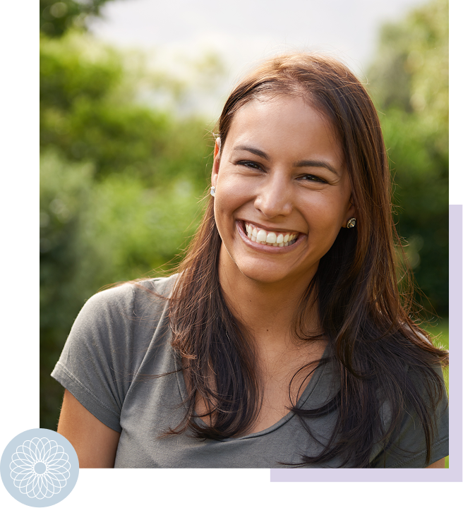 A young woman with long brown hair smiling outdoors with greenery in the background.