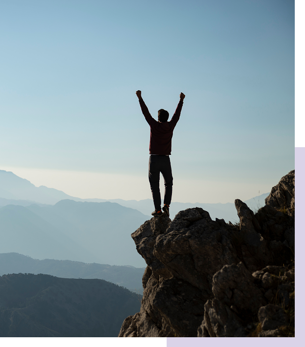 Person standing with arms raised on rocky mountain peak with mountain range in background during daytime.