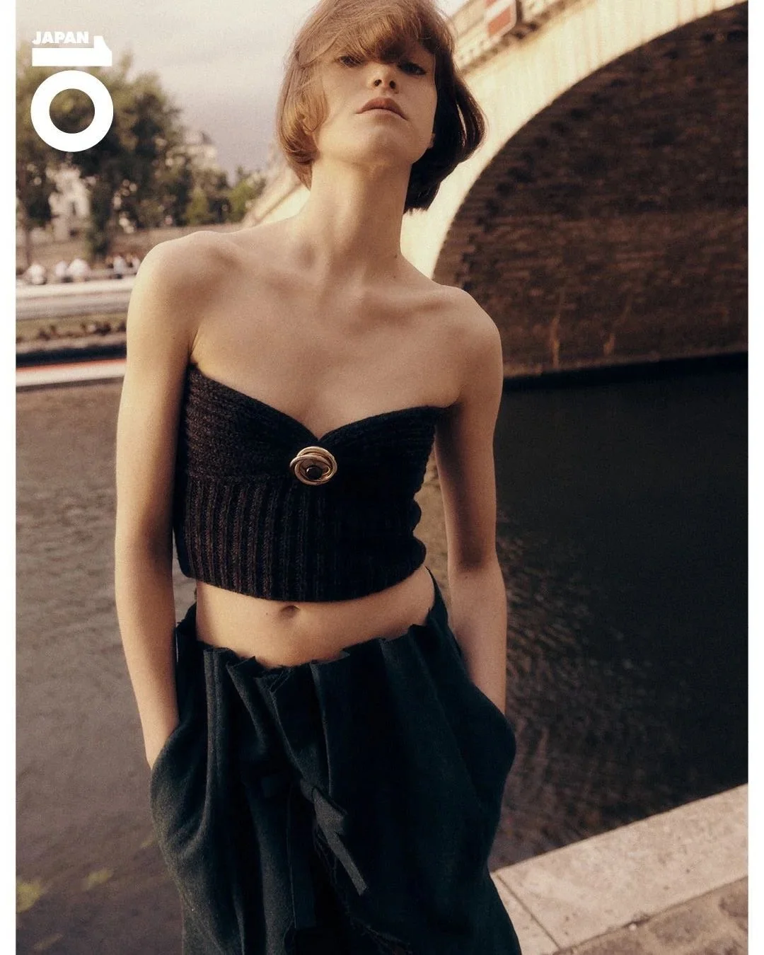 Jeune femme portant un haut noir court et un pantalon noir, se tenant près d'une rivière, sous un pont en briques, avec des arbres en arrière-plan.