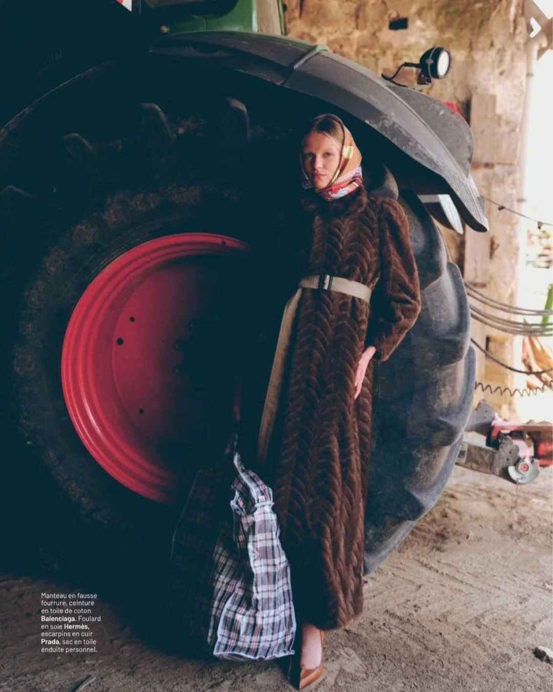 Une femme se tient à côté d'une grande roue de tractor en atelier. Elle porte un manteau en fausse fourrure marron, un foulard Hermes en soie, une ceinture, et un sac en toile à carreaux.