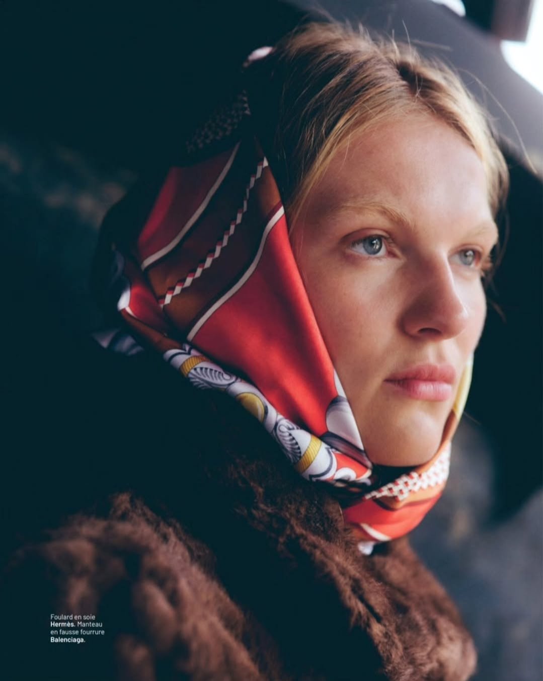 Une jeune femme porte un foulard en soie Hermès, un manteau en fausse fourrure Balenciaga, regardant pensivement.