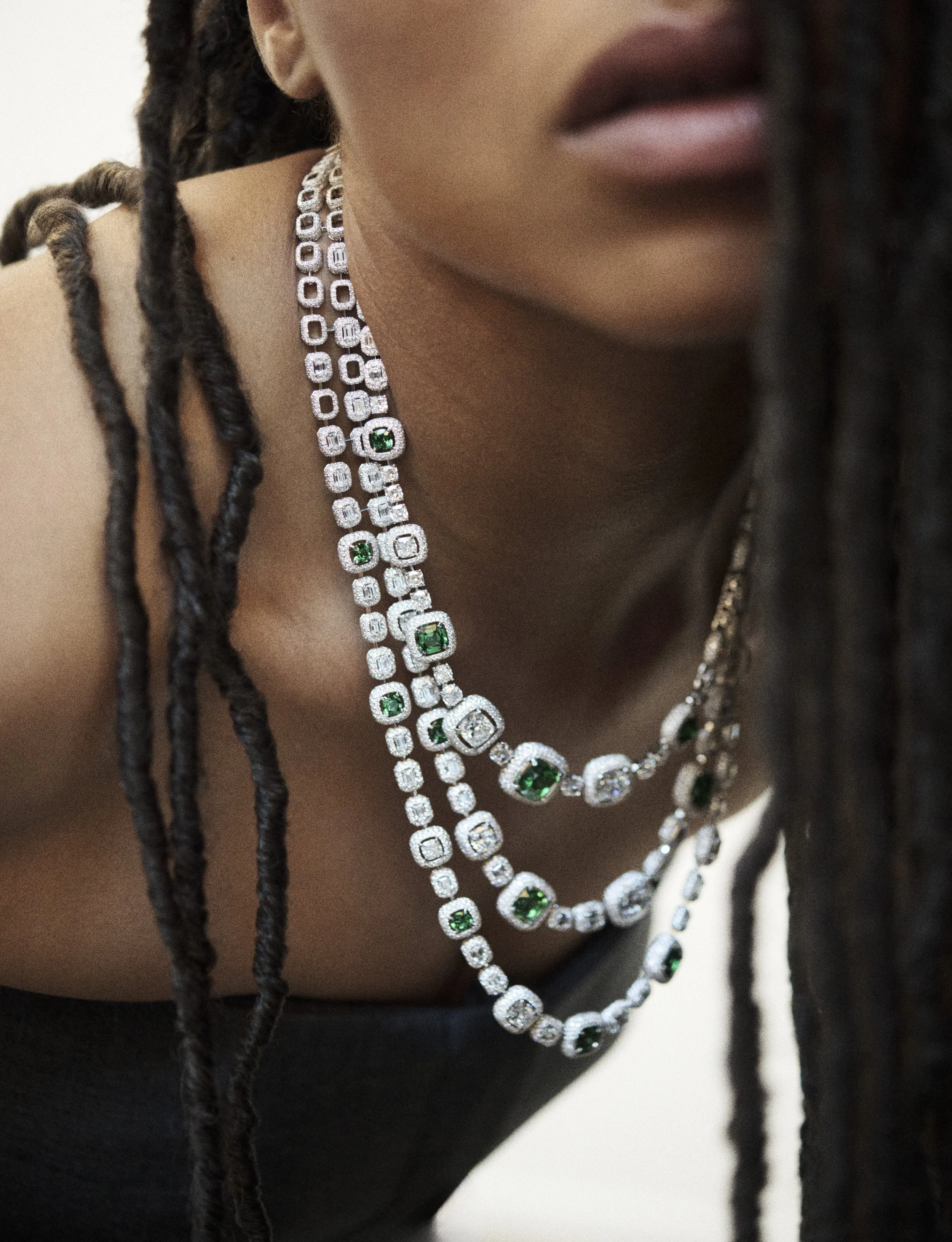Une personne porte plusieurs colliers ornés de diamants et d'émeraudes, avec ses dreadlocks encadrant le visage.
