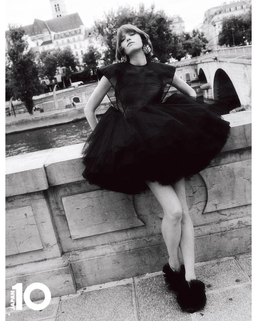 Une femme en robe noire tulle, posant sur un mur au bord de la rivière, avec des bâtiments et un pont en arrière-plan, en noir et blanc.
