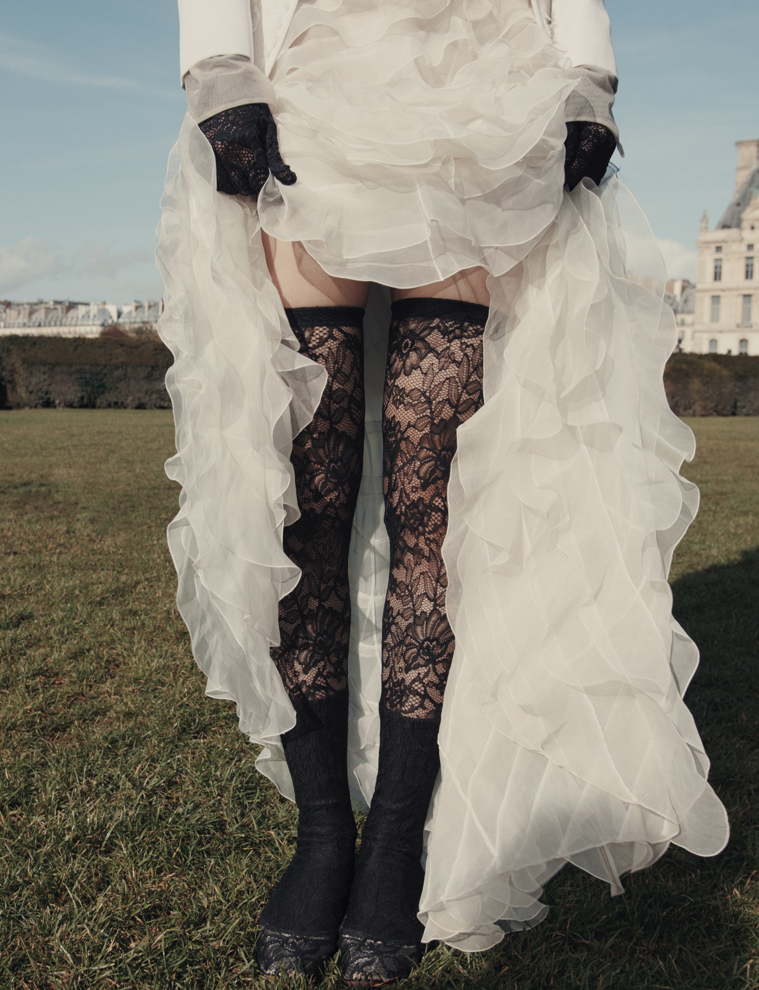 Une personne portant des chaussettes en dentelle noire, tenant une robe blanche ruffled à l'extérieur, avec un fond d'herbe et un bâtiment en arrière-plan.