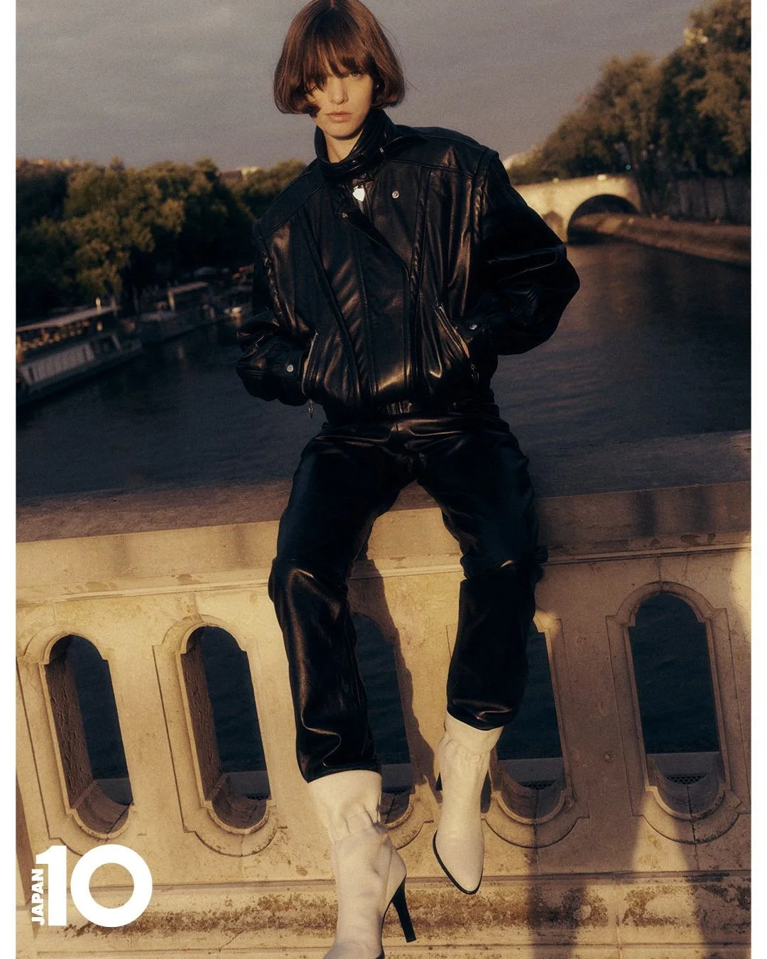 Une femme stylistique en veste en cuir noir et bottes blanches à talons, posant sur un pont au coucher du soleil avec une rivière en arrière-plan.