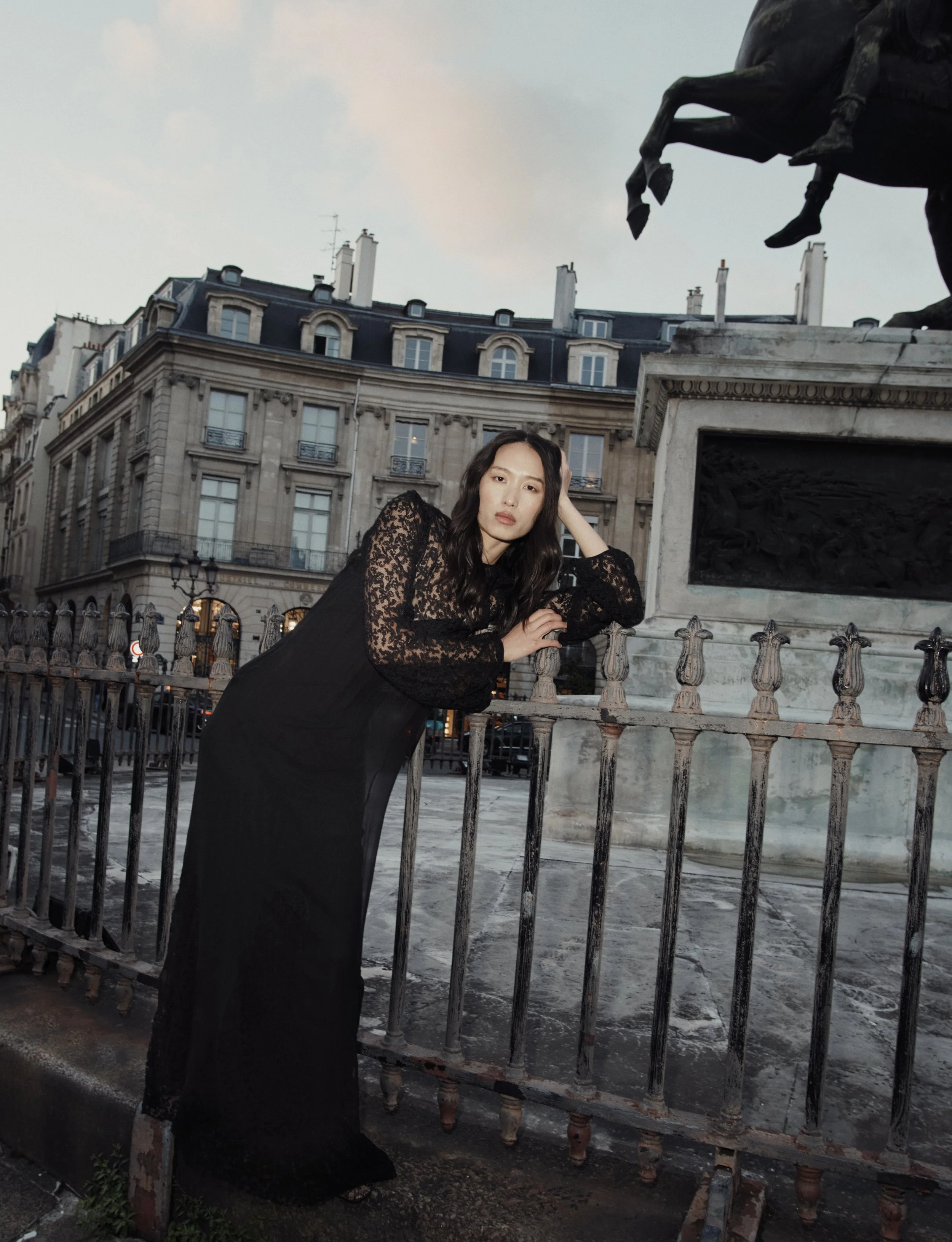 Une femme élégante en robe noire avec des manches en dentelle pose près d'une grille en fer forgé devant une statue équestre dans une ville européenne, probablement Paris, lors du crépuscule.