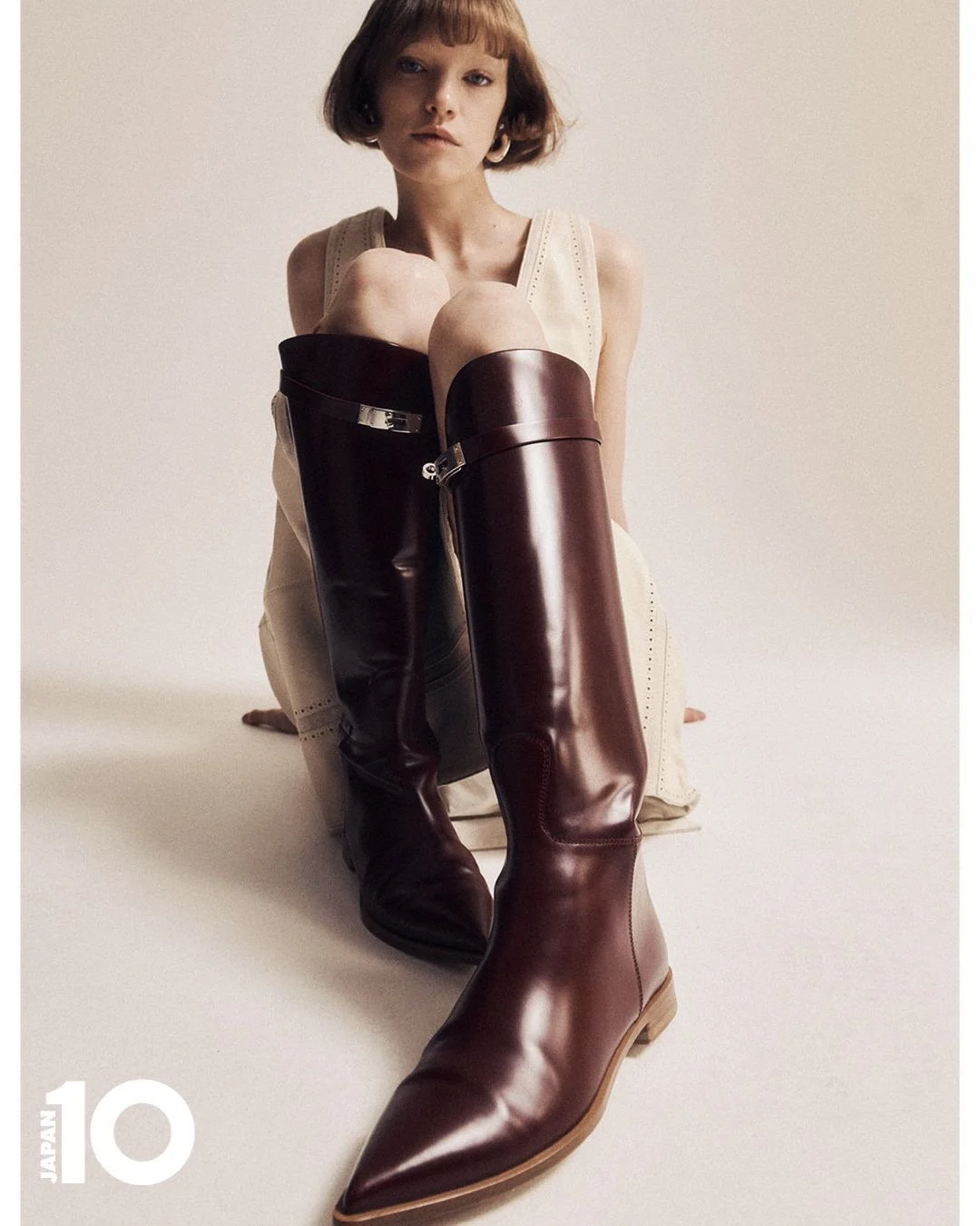 Une femme assise sur le sol portant une robe beige et des bottes en cuir de différentes couleurs, une noire et une marron.