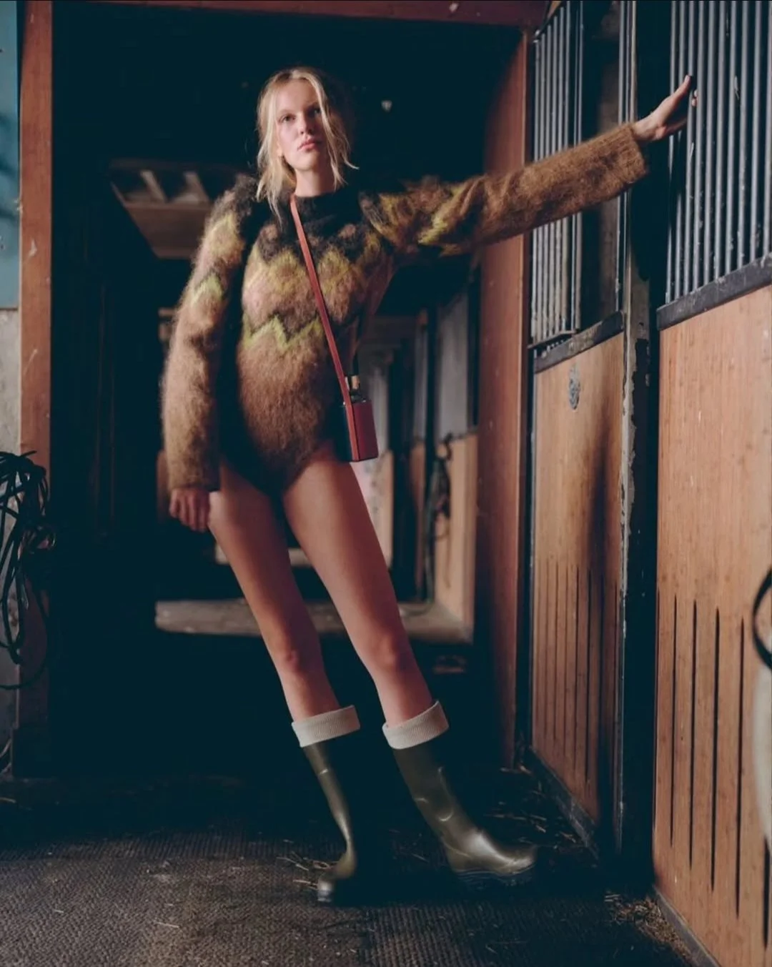 Une jeune femme avec de longues jambes, portant un pull en laine multicolore et des bottes en caoutchouc, dans une écurie, posant contre une barrière de box.