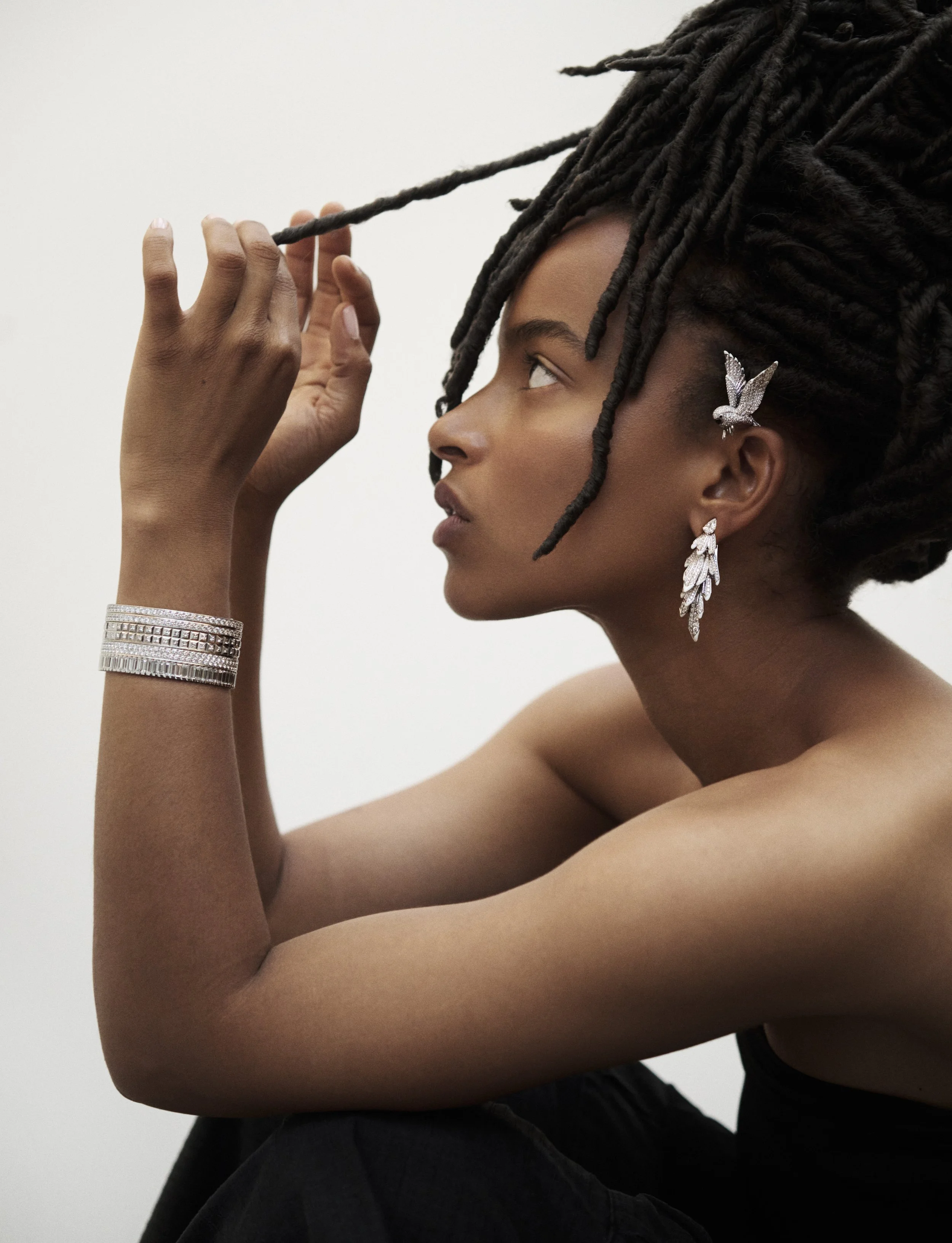 Femme avec dreadlocks portant des bijoux étincelants, regardant vers le haut, fond blanc.