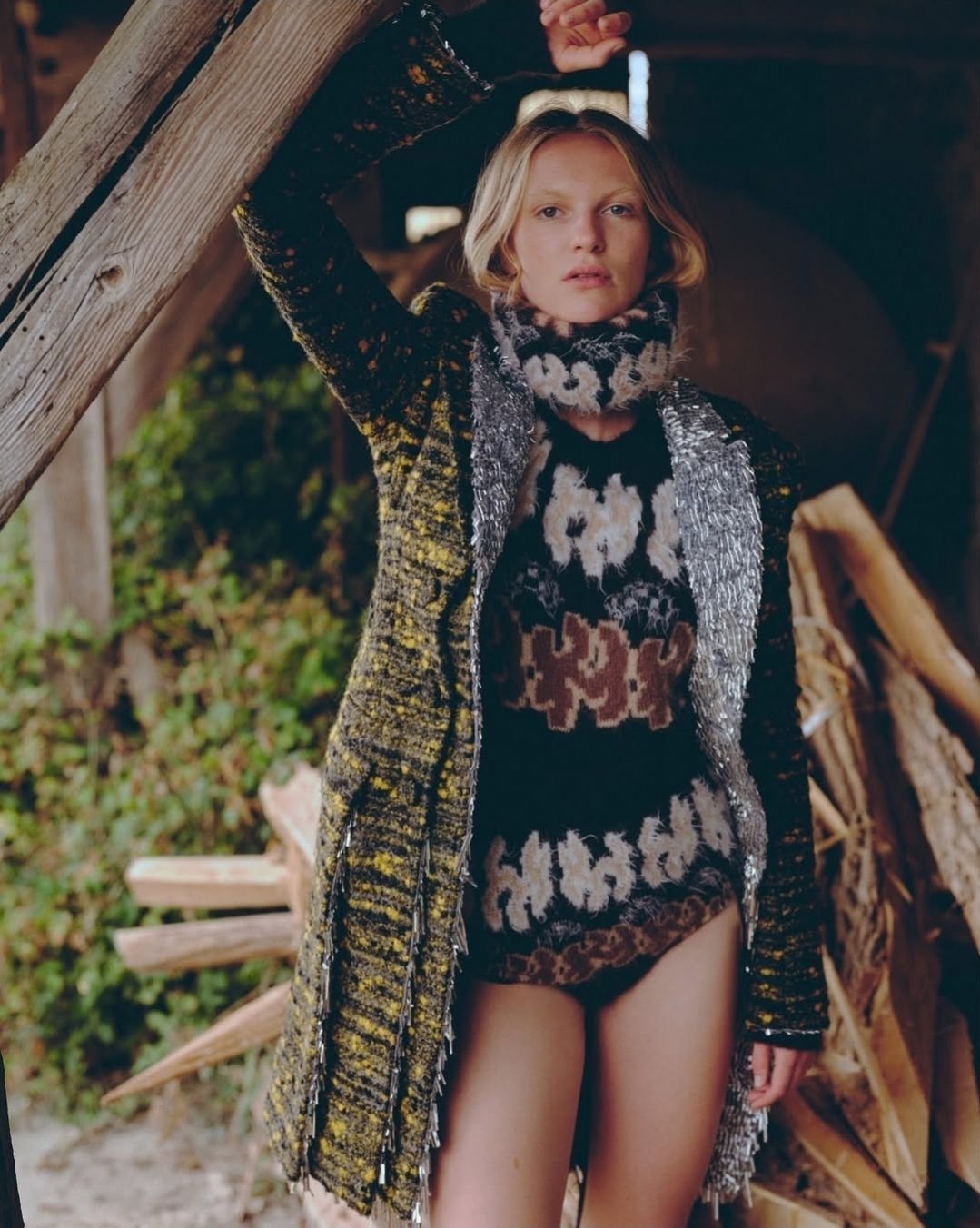 Jeune femme posant devant une structure en bois, portant un écharpe, un manteau orné de motifs ethniques, et des sous-vêtements, dans un environnement naturel.