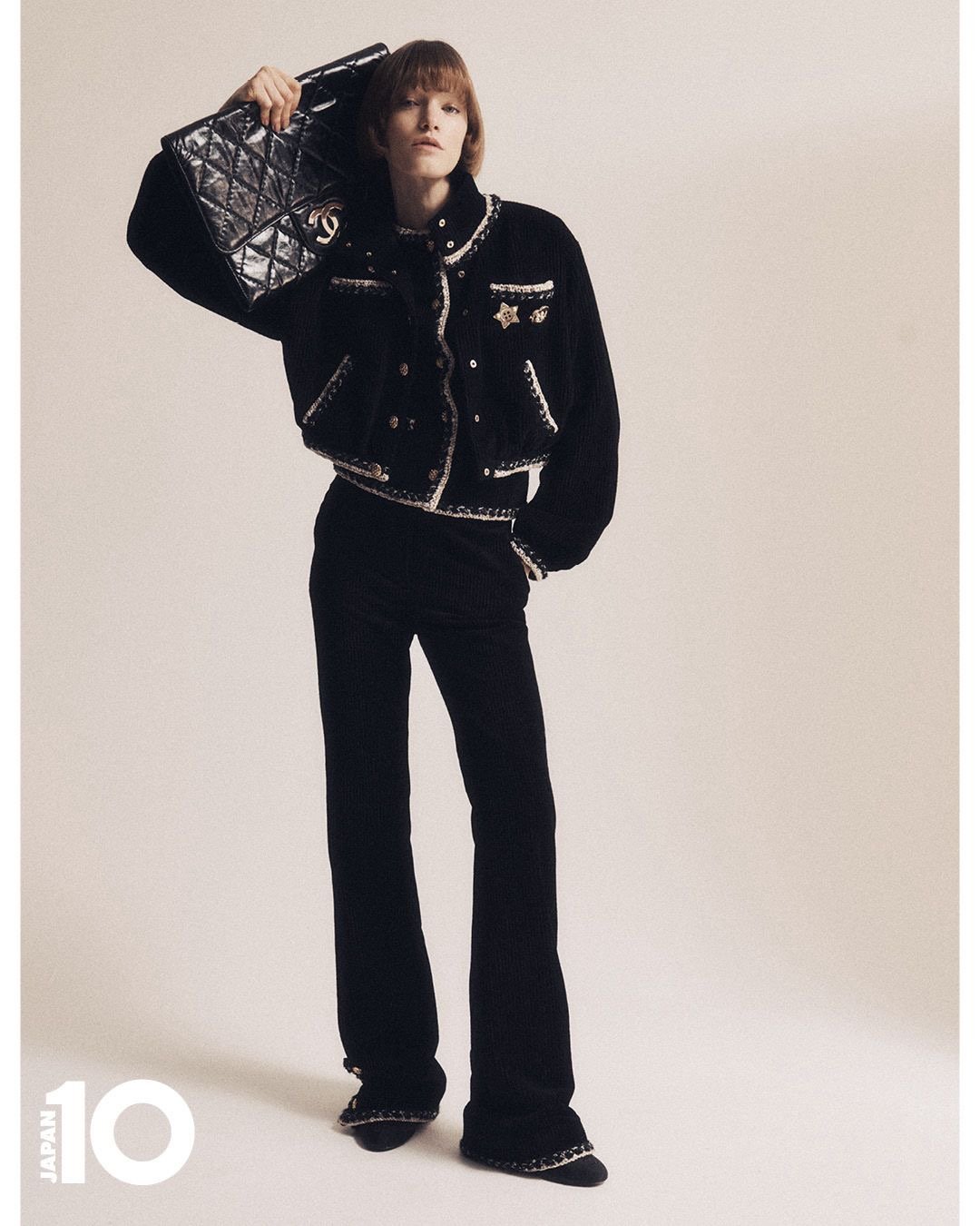 Une femme portant un pantalon et une veste en velours noir avec des détails en broderie blanche, tenant un sac à main noir quilté.