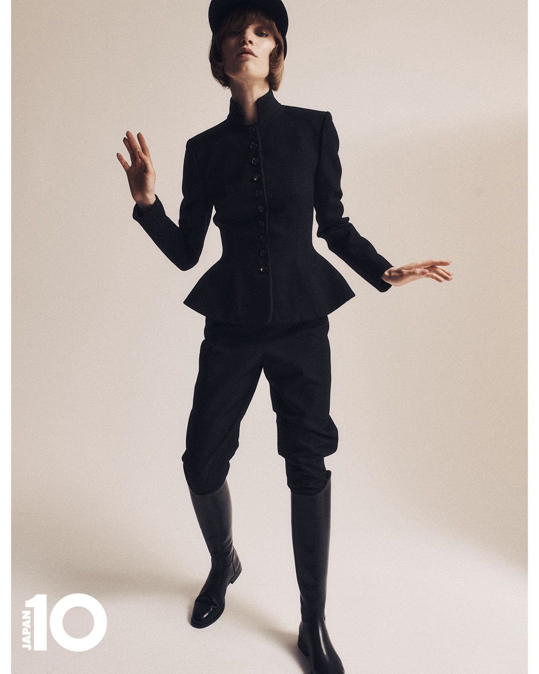 Une personne porte une tenue noire avec un chapeau noir, une veste cintrée et des bottes hautes, posant contre un fond neutre.