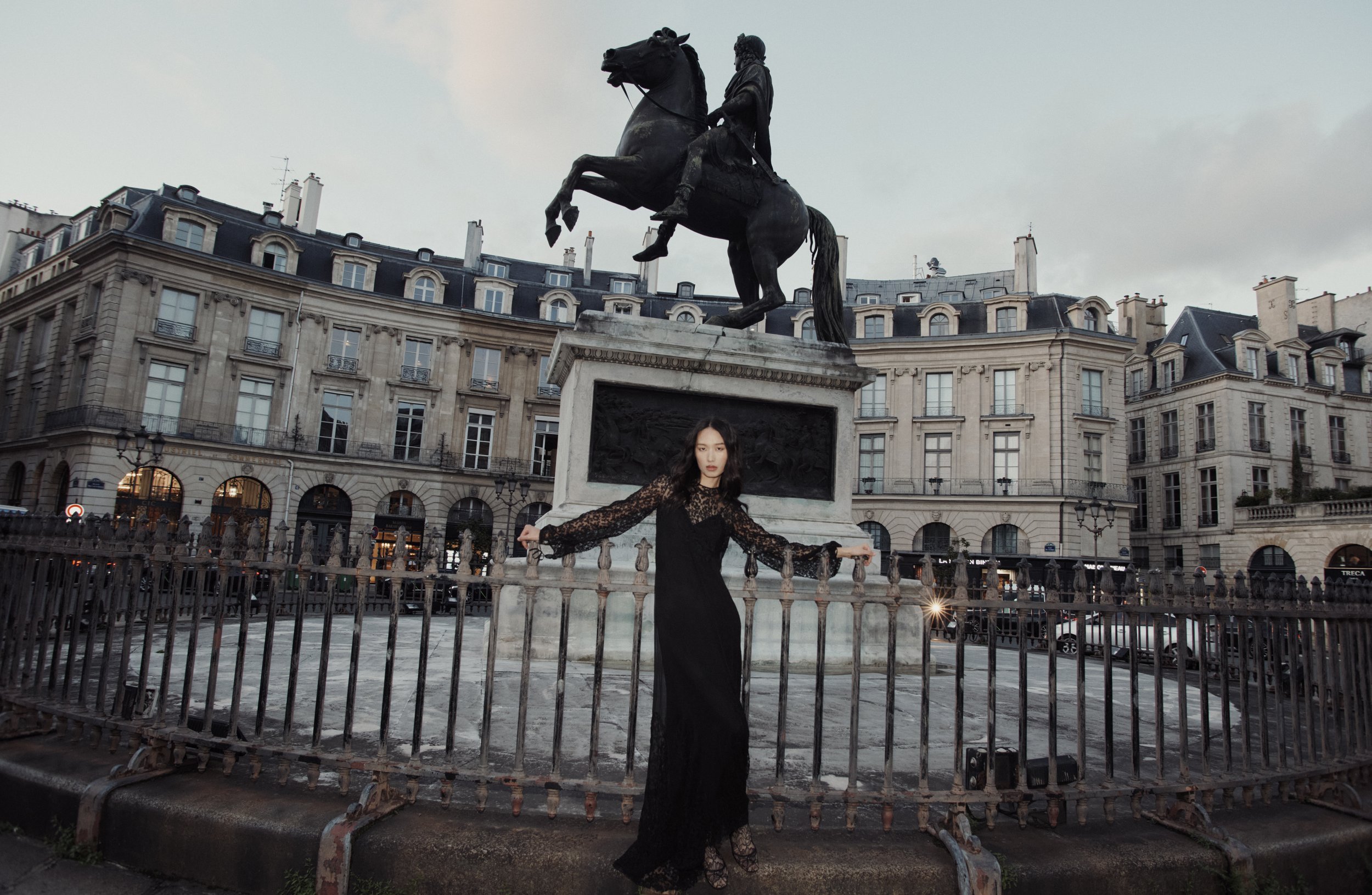 Une femme portant une robe noire en dentelle se tient devant une barrière en métal, devant une statue équestre en bronze représentant un homme à cheval sur une place urbaine. Des bâtiments classiques entourent la place, et il y a un ciel nuageux au-d