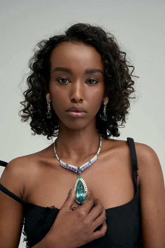 Une femme avec des cheveux bouclés porte des bijoux et une robe noire sans manches, regardant d'un air sérieux.