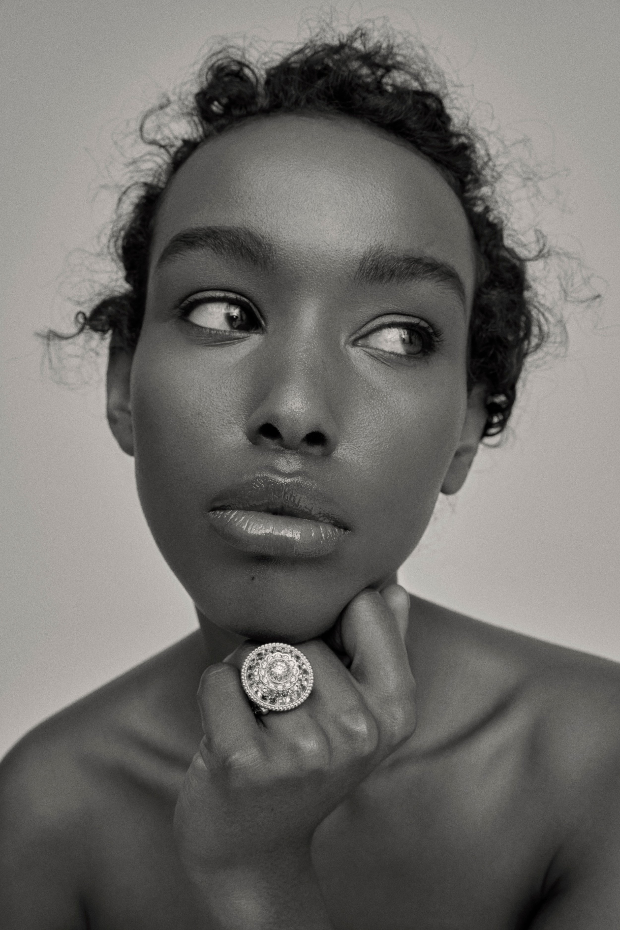 Portrait en noir et blanc d'une femme noire avec des cheveux bouclés courts, portant un grand anneau rétro avec des pierres, posant avec le doigt sous le menton, regardant de côté.