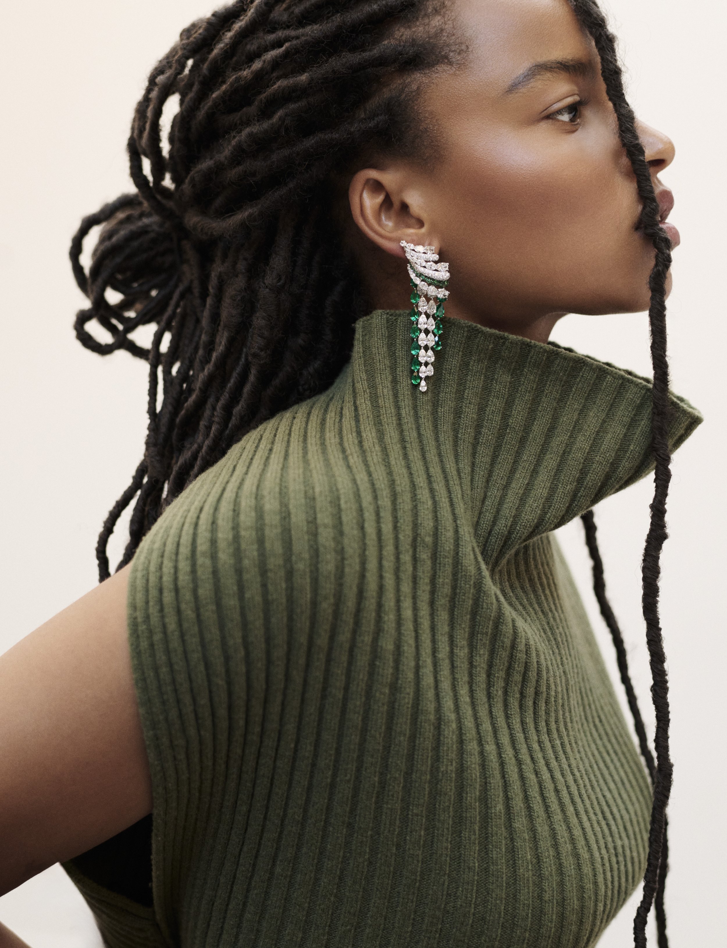Une femme avec des cheveux en dreadlocks porte une grande boucle d'oreilles orné de diamants et d'émeraudes et un pull vert à col haut en tissu côtelé.