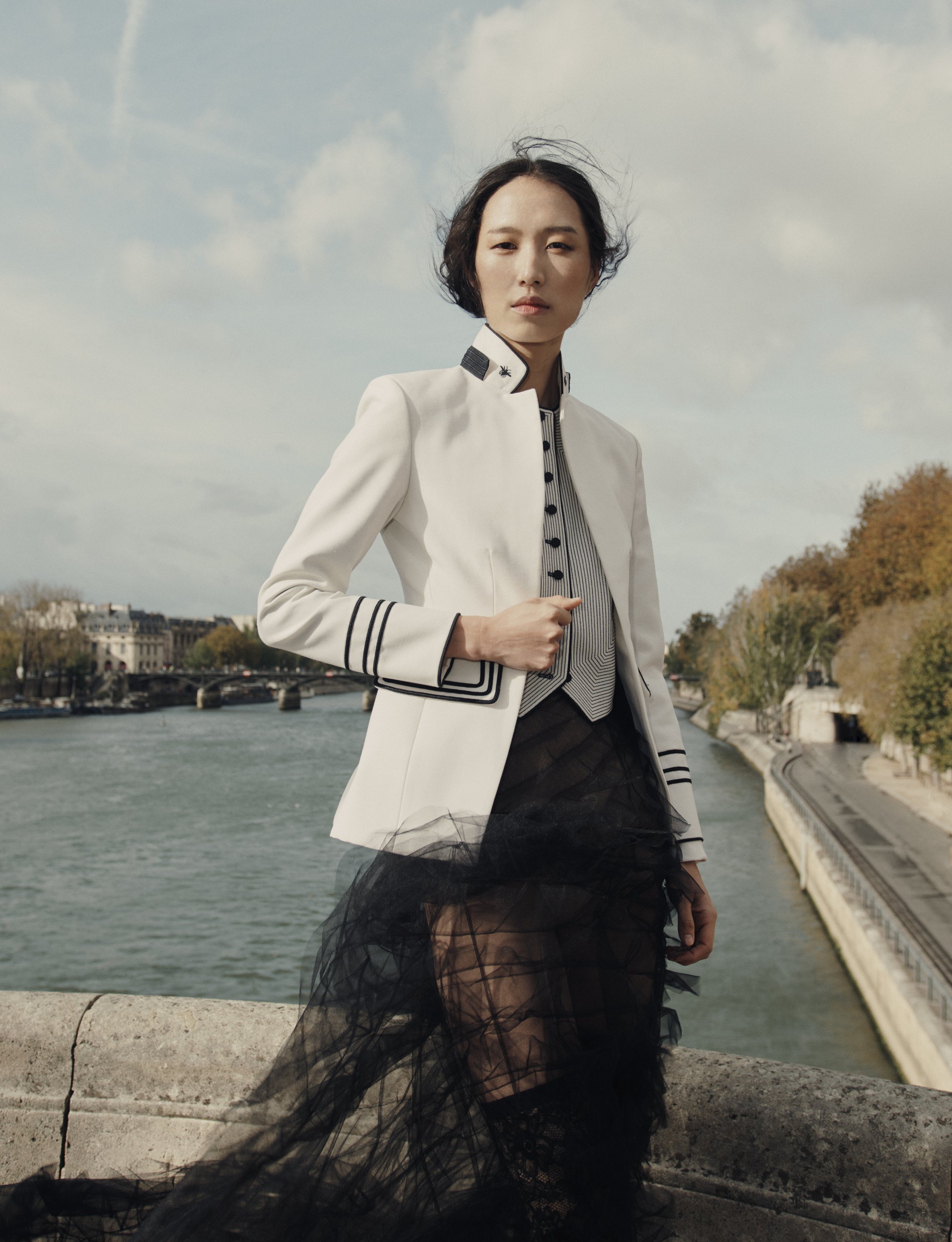 Femme posant près de la Seine à Paris, portant une tenue élégante avec une veste blanche, un haut à rayures et une jupe en tulle noir, sous un ciel nuageux.